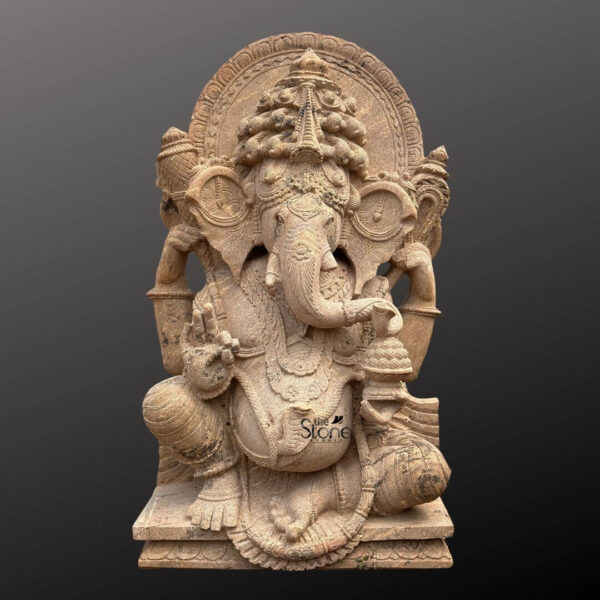 A beautifully detailed four-foot-tall sandstone Ganapathi stone vigraham or murti is depicted in a seated posture facing the viewer directly. The statue is carved from a single block of warm-toned sandstone exhibiting a rich texture with visible natural grain and variation. Lord Ganesha is seated on a modest tiered pedestal his left leg folded and his right foot resting on the base with the tip of his trunk elegantly curving to hold a modaka. His four arms are adorned with carved bracelets and armlets holding symbolic attributes a lotus bud in the upper right hand an axe or goad in the upper left a broken tusk in the lower right and the modaka bowl in the lower left. The Ganapathi vigraham is heavily adorned with intricate carvings including a prominent crown with elaborate filigree and floral motifs a sacred thread across his chest and multiple layers of detailed necklaces and chains. His large ears are fanned and decorated framing a serene and benevolent facial expression. The statue is backed by a richly carved circular prabhavali featuring a repeating leaf pattern and ornate decorative elements supported by pillars that emerge from behind Ganesha's shoulders. The entire sculpture rests upon a sturdy sandstone plinth which is minimally carved with a decorative base layer. This magnificent four-foot sandstone Ganesha statue is perfectly suited for installation as the central deity or a prominent shrine element within a temple. The scale of the murti combined with the exquisite craftsmanship and durable sandstone material makes it a revered focal point for worship and a testament to traditional Indian temple art. The warm natural color of the stone and the profound depth of the carving convey a sense of sanctity making it an ideal choice for temple environments seeking to enhance their spiritual ambiance and offer devotees a divine form for darshan. The intricate details and traditional iconography ensure it meets the spiritual and aesthetic requirements of a temple vigraham.