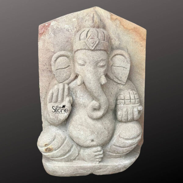 A three-quarter view studio photograph presents a monumental 36-inch tall abstract Ganesha statue masterfully hand-carved from a light-grey sandstone with an aged antique patina. The sculpture depicts the Hindu deity Ganesha in a seated posture characterized by a simplified yet profound form that emphasizes mass and spiritual presence over intricate detail. The stone exhibits a naturally textured slightly porous surface with warm earthy ochre and reddish-brown mineral deposits embedded within the crevices enhancing its ancient weathered appearance. The statue retains the overall rectangular block shape of the original stone from which the deity's form is carved in deep relief giving it a raw powerful presence. Ganesha is shown with his characteristic elephant head which is depicted with closed contemplative eyes and a gently curved trunk resting to the left. His large fan-like ears are flat and minimally adorned merging into a simple conical crown with a carved floral medallion. The sculpture has four arms in a stylized arrangement: the upper right hand holds a modak the upper left holds a parashu the lower right is in Abhaya mudra and the lower left holds a pasha. The deity's corpulent belly is centrally prominent delineated by a simple sacred thread and a carved belt while his legs are folded in a meditative half-lotus position. The abstract nature of the carving style is evident in the broad unadorned planes of the body and limbs and the lack of elaborate jewelry which focuses the viewer on the fundamental archetypal form of the deity. The statue is carved from a solid heavy piece of sandstone with the top edge of the block left naturally uneven and rugged further contributing to its archaic temple-artifact aesthetic. The sandstone material with its natural color variations and textures makes this large Ganesha murti a striking piece of spiritual art suitable for both traditional and modern spaces seeking a touch of ancient calm.