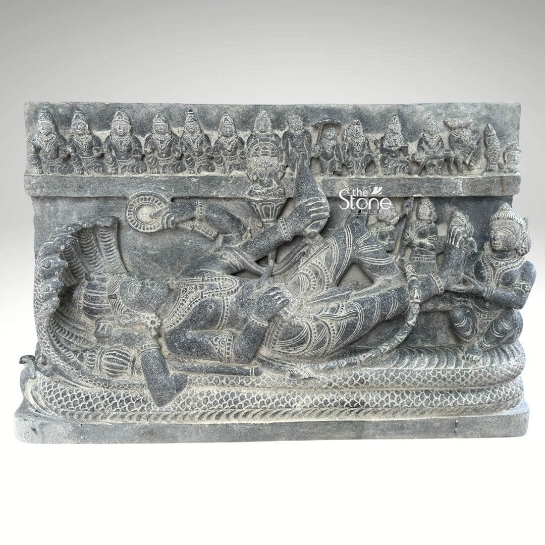 A finely carved blackstone relief panel measuring approximately 30 inches depicting the reclining form of Lord Vishnu known as Anantashayana. The central figure of Vishnu is shown in a state of cosmic sleep resting peacefully upon the multi-hooded serpent Shesha or Ananta whose coils form a textured bed and whose seven hooded heads fan out to create a protective canopy above the deity's head. Vishnu is adorned with traditional jewelry a crown and elaborate garments with his right hand gently supporting his head. The sculpture includes several attendant figures carved in high relief. At Vishnu's feet his consort Goddess Lakshmi is seated and shown massaging his foot a gesture of devotion. Below the main reclining figure the detailed coils of the serpent are intricately rendered. At the top of the stele a frieze of miniature figures representing the ten avataras of Vishnu is carved horizontally across the panel adding a comprehensive narrative element to the divine iconography. The entire murti is carved from a single block of deep rich black stone exhibiting a matte tactile finish that emphasizes the detailed carvings and high relief. The stone's texture is palpable conveying a sense of ancient artistry and reverence. The sculpture is presented against a plain neutral grey background which isolates the form and draws the viewer's eye directly to the complex narrative and divine iconography depicted in the stone. This piece is an exemplary representation of traditional Indian temple art.