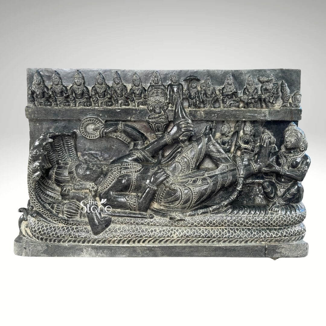 A detailed Anantashayana Vishnu Antique Stone Sculpture approximately 30 inches tall depicting the reclining form of Lord Vishnu in his cosmic slumber. The sculpture is carved from a single block of rich blackstone showcasing a deep matte texture that highlights the intricate high-relief carvings. The central figure of Vishnu is shown resting upon the multi-hooded serpent Shesha whose coils form a textured bed beneath him and whose seven hooded heads fan out to create a protective canopy above the deity's crowned head. Vishnu is adorned in traditional regalia including a kiritamukuta elaborate necklaces armlets waistbands and flowing lower garments all rendered with fine detail in the stone. His right hand gently supports his head, while his left hand rests along his body holding a conch. The sculpture includes several attendant figures carved in relief. Seated at Vishnu's feet is his consort Goddess Lakshmi who is shown massaging his foot with devotion. Above the main reclining figure a frieze of miniature figures representing the ten avataras of Vishnu is carved horizontally adding a comprehensive narrative to the divine iconography. The entire stele is carved from a deep rich black stone exhibiting a matte tactile finish that emphasizes the high relief and detailed craftsmanship. The natural variations in the stone's color and texture convey a sense of age and authenticity consistent with an antique piece. The sculpture is presented against a plain neutral grey background which isolates the form and draws the viewer's eye directly to the complex narrative and divine iconography depicted in the stone. This detailed carving serves as an exemplary illustration for an article on Anantashayana Vishnu antique stone sculptures.