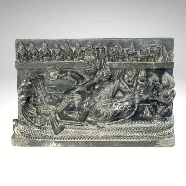 A detailed black stone relief carving, approximately 30 inches in height depicting the reclining form of Lord Vishnu in the Anantashayana pose. The central figure of Vishnu is shown resting peacefully upon the multi-hooded serpent Shesha who forms a protective canopy above his head with his seven hooded heads. Vishnu is adorned with traditional jewelry a crown and elaborate garments with his right hand gently supporting his head while his left hand rests along his body. The composition includes several attendant figures carved into the stele. Seated at Vishnu's feet is his consort Goddess Lakshmi who is shown massaging his foot. Above the serpent canopy a frieze of miniature figures representing the ten avataras of Vishnu is carved horizontally. Below the main reclining figure the serpent's coils are intricately detailed and a small offering stand with ritual items is also visible. The entire sculpture is carved from a single block of deep rich black stone showcasing a matte finish that highlights the intricate detailing and high relief of the carving. The stone's texture is palpable conveying a sense of ancient artistry and reverence. The statue is presented against a plain neutral grey background which isolates the form and draws the viewer's eye directly to the complex narrative and divine iconography depicted in the stone.