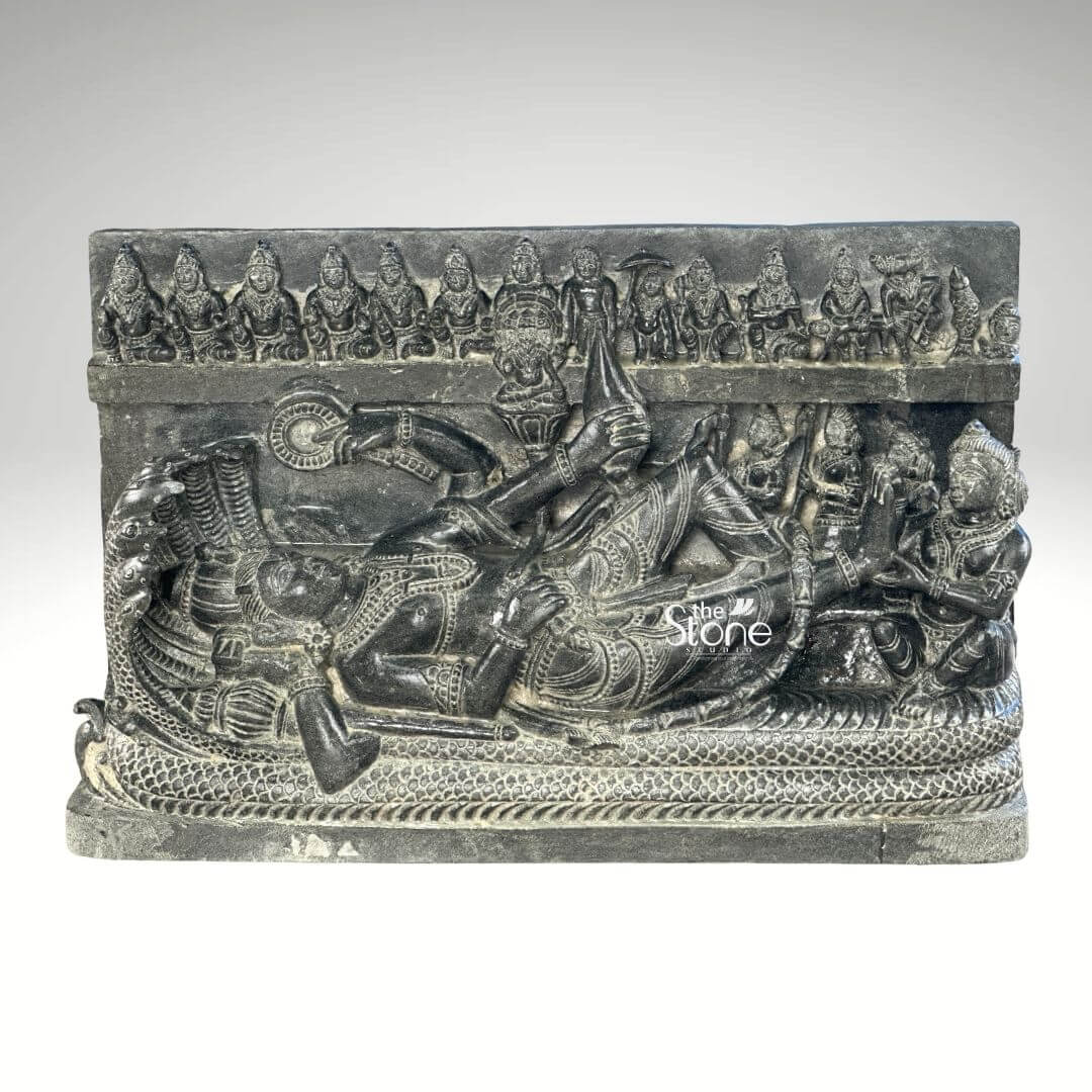 A detailed black stone relief carving, approximately 30 inches in height depicting the reclining form of Lord Vishnu in the Anantashayana pose. The central figure of Vishnu is shown resting peacefully upon the multi-hooded serpent Shesha who forms a protective canopy above his head with his seven hooded heads. Vishnu is adorned with traditional jewelry a crown and elaborate garments with his right hand gently supporting his head while his left hand rests along his body. The composition includes several attendant figures carved into the stele. Seated at Vishnu's feet is his consort Goddess Lakshmi who is shown massaging his foot. Above the serpent canopy a frieze of miniature figures representing the ten avataras of Vishnu is carved horizontally. Below the main reclining figure the serpent's coils are intricately detailed and a small offering stand with ritual items is also visible. The entire sculpture is carved from a single block of deep rich black stone showcasing a matte finish that highlights the intricate detailing and high relief of the carving. The stone's texture is palpable conveying a sense of ancient artistry and reverence. The statue is presented against a plain neutral grey background which isolates the form and draws the viewer's eye directly to the complex narrative and divine iconography depicted in the stone.