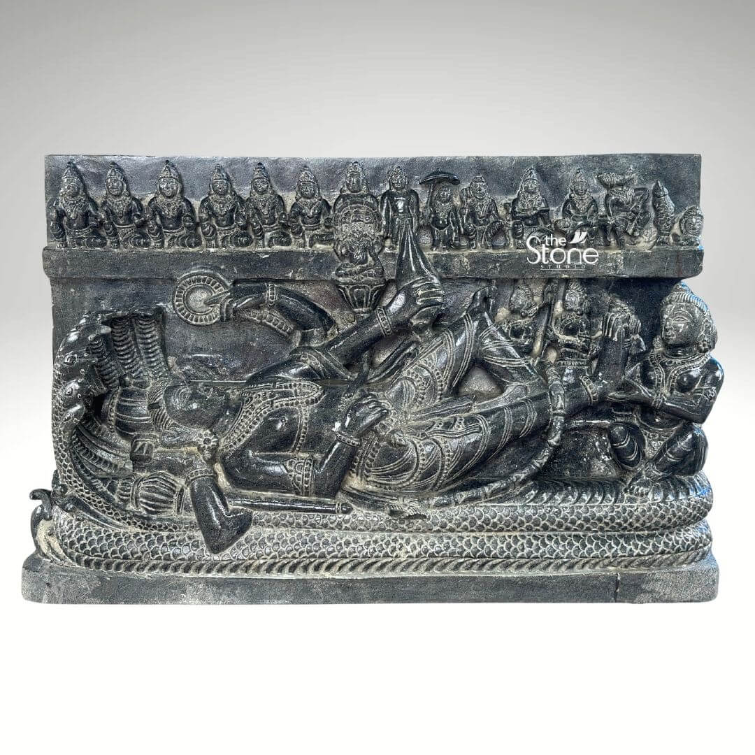 A deeply carved black stone statue approximately 30 inches in height depicting the reclining form of Lord Vishnu known as Ananthasayana Perumal. The central figure of Vishnu is shown resting peacefully upon the coils of the multi-hooded serpent Shesha who forms a protective canopy above the deity's head with his seven hooded heads. Vishnu is adorned with traditional jewelry a tall crown and elaborate garments with his right hand gently supporting his head and his left arm resting along his body holding a conch shell. The sculpture includes several attendant figures carved into the stele. Seated at Vishnu's feet is his consort Goddess Lakshmi who is shown massaging his foot. A smaller figure perhaps the sage Markandeya is also depicted near his head. Above the serpent canopy a frieze of miniature figures representing the ten avataras of Vishnu is carved horizontally across the panel adding a comprehensive narrative to the divine iconography. Below the main reclining figure the serpent's coils are intricately detailed along with a small offering stand with ritual items. The entire murti is carved from a single block of deep rich black stone showcasing a matte finish that highlights the intricate detailing and high relief of the carving. The stone's texture is palpable conveying a sense of ancient artistry and reverence. The statue is presented against a plain neutral grey background, which isolates the form and draws the viewer's eye directly to the complex narrative and divine iconography depicted in the stone. This detailed carving is an exemplary representation of traditional South Indian temple art making it a perfect illustration for an article discussing Ananthasayana Perumal stone statues.