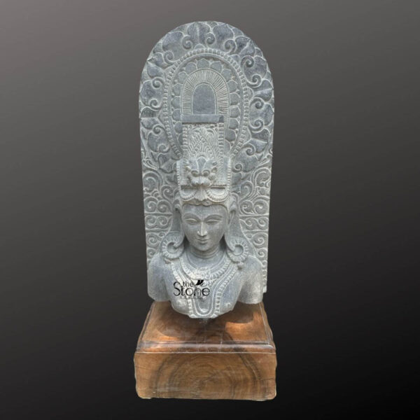 A twenty-three-inch-tall antique Lord Shiva stone bust statue is depicted frontally mounted on a simple dark wooden block pedestal. The sculpture is meticulously carved from a single block of grey stone exhibiting a rich aged texture with natural variations and a weathered patina that underscores its antiquity. Lord Shiva is portrayed with a serene contemplative facial expression with eyes gently closed in meditation arched eyebrows, and a subtle compassionate smile. His hair is intricately styled in a traditional jata forming a high crown adorned with delicate carvings of a crescent moon, a serpent, and a central floral motif with further details of coiled locks falling onto his shoulders. The bust features elaborate ornamentation including large circular earrings and multiple layered necklaces with intricate pendants that rest upon his chest all hand-carved with precision. A sacred thread is visible across his torso. Behind the deity's head a richly carved ornate prabhavali rises featuring detailed symmetrical floral and scrollwork patterns that frame his divine form. The craftsmanship showcases the traditional South Indian temple art style with deep undercuts and high relief that create a profound sense of depth and reverence. The entire sculpture rests securely on a dark polished wooden plinth providing a stable base. This magnificent twenty-three-inch antique Lord Shiva stone bust statue is a testament to the timeless devotion and artistic skill of ancient Indian craftsmen. The detailed iconography from the serene meditative face to the intricate jewelry and the elaborate prabhavali makes it a revered centerpiece for any spiritual collection or home altar. The durable stone material and the profound expression of the sculpture make it a lasting symbol of divine wisdom meditation and cosmic energy perfectly suited for those seeking a high-quality antique Shiva murti to enhance their sacred space. The natural color of the stone and the profound depth of the carving convey a sense of sanctity and spiritual presence.
