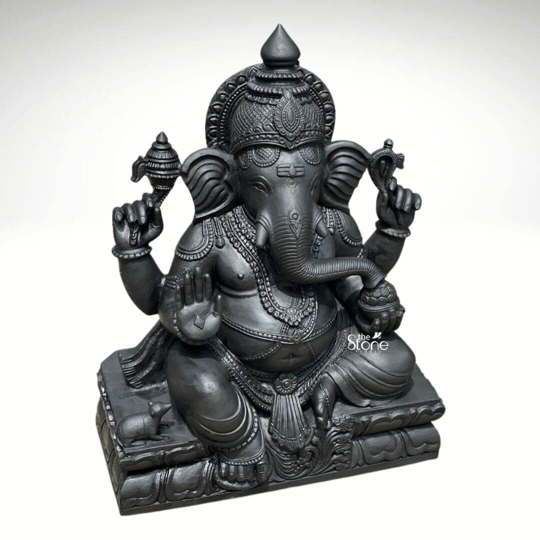 A magnificent 2.5-foot-tall black stone Ganapathi statue stands as a profound example of traditional South Indian sculptural art perfectly scaled for both home and temple environments. Hand-carved from a single block of deep black soapstone the statue depicts the Hindu deity Ganesha in a traditional seated posture known as the Lalitasana pose with his right leg folded and left leg slightly raised. The dark polished stone surface exhibits a subtle cool sheen under the studio lighting highlighting the smooth texture of the material. The artisan has lavished attention on the intricate details from the ornate crown and multiple necklaces adorning his form to the delicate folds of his flowing dhoti and the individual petals of the lotus throne he occupies. Ganesha is depicted with four arms each holding a symbolic item with great precision his upper right hand holds an ankusha his upper left holds a pasha and his lower right hand is in the Abhaya Mudra bestowing blessings upon devotees. His lower left hand holds a modak resting in his lap with his trunk gently curled to sample the offering. The statue features a prominent finely carved prabhavali behind the deity adorned with traditional patterns and culminating in a kirtimukha at the apex. His loyal vahana the mouse is seated attentively at his feet to the left completing the divine composition. This black stone Ganapathi statue strikes an ideal balance being large enough to serve as a central deity in a home puja room or a smaller temple sanctum yet detailed enough to be appreciated as a significant decorative piece. The substantial weight and durable stone material ensure its longevity making it a lasting investment for spiritual practice or cultural decor. The clean studio photograph against a plain white background emphasizes the statue's three-dimensional quality and the artisan's meticulous work on the serene facial expression and decorative elements perfectly illustrating the grace and cultural richness that a well-crafted black stone Ganesha statue brings to any consecrated space.