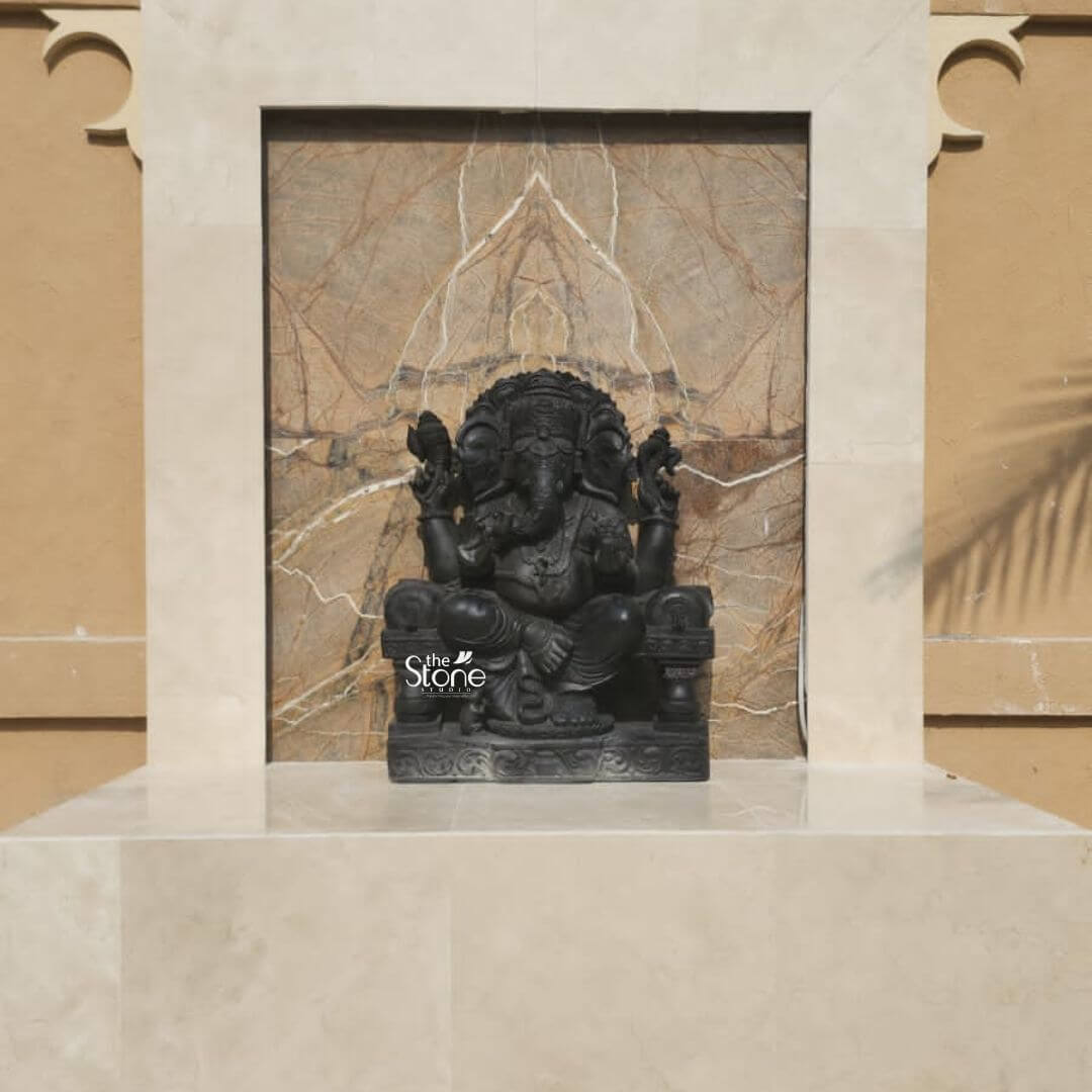 A three-quarter view photograph captures a magnificent Black Stone Ganesha Statue meticulously hand-carved from a single piece of deep dark basalt. Standing approximately 3 feet tall this imposing statue depicts Lord Ganesha in a traditional seated posture known as lalitasana with his right leg folded and his left leg bent. The statue's smooth matte finish accentuates the natural rich color of the stone and the exceptional precision of the sculptural details. The deity is adorned with elaborate deeply-cut ornamentation across his form including multiple necklaces armbands wristlets and anklets all rendered in the durable black stone. He wears a richly carved crown and a sacred thread across his chest. He has four arms his upper right hand holds a parashu and his upper left holds a pasha. His lower right hand is raised in the abhaya mudra while his lower left hand cradles a modaka which his gracefully curved trunk is poised to taste. This Black Stone Ganesha statue is elegantly enshrined within a custom-built recessed niche on a warm textured exterior wall showcasing its suitability for a home shrine or garden setting in Dubai. The niche features a simple frame of polished cream-colored stone and a backdrop of textured beige marble with subtle white veining which provides a beautiful contrast to the dark stone of the statue. The entire composition sits on a substantial polished marble shelf emphasizing its revered placement within a contemporary Dubai architectural context.