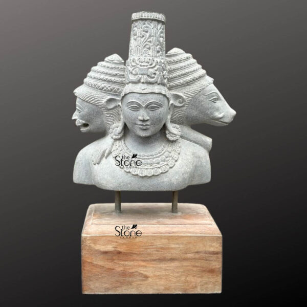 A detailed 19-inch bust sculpture of the Hindu Trimurti meticulously hand-carved from a single piece of grey sandstone. The central figure represents Lord Vishnu facing forward with a serene expression and traditional ornate crown. To his left is the fierce bearded face of Lord Mahesh and to his right is the calm bovine visage of Lord Brahma symbolizing the unity of creation preservation and destruction. The three heads are conjoined at the neck and shoulders creating a cohesive tri-faced form. Each face is distinguished by specific features and expressions showcasing the artisan's skill in capturing the distinct personalities of the deities. The sculpture is adorned with traditional carved jewelry including multiple strands of necklaces that drape across the chest of each figure enhancing the divine aesthetic. The stone bust is mounted on two metal rods fixed into a solid rustic rectangular wooden block base providing a stable and elegant presentation. The natural granular texture of the grey sandstone contrasts beautifully with the smooth warm grain of the wood. The sculpture is set against a simple neutral grey background which isolates the piece and draws the viewer's eye directly to the intricate details of the Tridev heads. This high-quality image is an excellent representation of classical Indian stone artistry making it an ideal illustration for an article about Brahma Vishnu Mahesh stone statues.