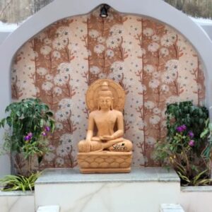A detailed eye-level photograph capturing a beautiful 2.5ft Buddha statue hand-carved from warm brown sandstone. The statue is depicted in a traditional seated posture with the left hand resting in the lap and the right hand touching the earth. It is seated upon a multi-tiered pedestal also carved from matching brown sandstone with a decorative lotus petal motif. The stone's natural grain and warm earthy tones are clearly visible highlighting the artisan's skillful carving on the robe's folds facial features and the ushnisha.The Buddha statue is positioned within a tranquil outdoor garden setting set against a dramatic backdrop. It sits on a raised white marble altar which provides a clean contrast to the warm brown stone. Behind the altar there is an arched stone alcove. The wall within this alcove is decorated with exquisite textured sandstone panels featuring deeply carved repeating patterns of white flowering branches and leaves all rendered in natural sandstone shades. A small focused spotlight above the arch illuminates the statue from the front casting soft defined shadows that emphasize the depth and serenity of the form.Flanking the marble altar are two healthy green plants with small purple flowers adding a touch of natural life and color to the devotional space. The ground around the altar is paved with natural stone tiles and further soft green foliage is visible on the periphery creating a sense of peace and harmony. The entire composition conveys a profound sense of tranquility and spiritual connection making this Brown Sandstone Buddha 2.5ft Statue a focal point of meditation and reverence. The careful integration of the statue with its natural surroundings and the detailed background carving reflects a high level of craftsmanship and artistic sensibility.