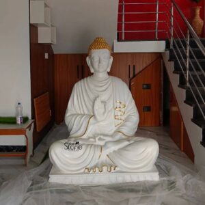 A five-foot-tall seated Buddha statue finished in pristine white with opulent gold accents stands as the serene centerpiece within an indoor living space in Ponneri Tamil Nadu. Carved from durable fibreglass the statue exhibits a smooth matte white surface on its body and robes which is elegantly contrasted by a gleaming metallic gold finish on its intricately curled ushnisha elongated earlobe and the delicate sanghati border that drapes over the left shoulder. Lord Buddha is depicted in a traditional meditative posture seated upon a multi-tiered lotus pedestal with his legs crossed in the vajra position and his hands held in the gesture of fearlessness and reassurance the abhaya mudra. His right hand is raised with the palm facing outward while his left hand rests gently on his lap. The statue is positioned directly on the polished marble floor which is partially covered by protective bubble wrap near the base. The background reveals a contemporary home interior with a modern staircase featuring stainless steel railings and dark stone steps rising on the right. Behind the Buddha a warm wooden-paneled wall panel adds a touch of natural texture flanked by a plain white wall. To the left a simple console table holds a water bottle and a small patch of artificial grass. The scene is illuminated by natural daylight filtering in from an unseen source highlighting the clean lines radiant gold details and serene benevolent facial expression of the Buddha. This magnificent four-foot fibreglass Buddha statue is an exemplary piece of contemporary temple art expertly crafted to bring peace and tranquility to any home or institution in Ponneri. The durable fibreglass material ensures the sculpture's longevity and lightweight nature making it a practical and elegant choice for interior decoration. The fusion of classic Buddhist iconography with a modern white and gold finish creates a powerful spiritual presence making this murti a revered focal point for meditation prayer or as a profound symbol of mindfulness within the cultural landscape of Tamil Nadu. The exquisite detailing and serene posture ensure it meets the highest aesthetic and spiritual requirements for a Buddha statue.