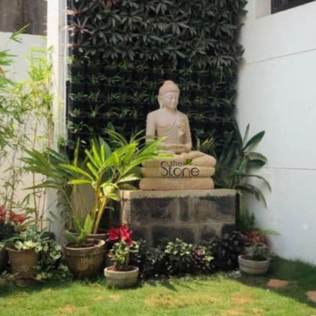 A three-foot-tall seated Buddha statue meticulously carved from warm light-colored sandstone forms the spiritual center of this serene terrace garden. The statue is positioned in a meditative Dhyana Mudra pose with hands resting gently in the lap. It is placed upon a weathered rough-hewn stone plinth giving it a grounded ancient presence. The natural texture of the sandstone is highlighted by the soft natural daylight showcasing the artisan's detailed work on the serene facial expression and the folds of the robe. The garden setting surrounding the Buddha is designed to create a peaceful sanctuary on a rooftop. Directly behind the statue rises a tall lush vertical living wall completely covered in dense dark green foliage. This wall serves as a rich textured backdrop that enhances the statue's form. The base of the plinth is surrounded by a collection of various potted plants including large palms ferns and smaller leafy plants with touches of deep red and purple adding a vibrant layer of nature. To the left a stand of tall bamboo stalks adds verticality and a distinctly Asian-inspired aesthetic contributing to the tranquil atmosphere. This image perfectly illustrates a harmonious integration of garden art into a modern urban outdoor space. The three-foot sandstone Buddha is an ideal scale for a terrace garden in Hubli providing a focal point that balances the living elements with a sense of calm. The surrounding plant palette featuring both tropical and resilient foliage thrives in the environment while the statue’s durable sandstone material is well-suited for outdoor display. The entire corner composition offers inspiration for creating a contemplative retreat demonstrating how a carefully chosen stone statue can elevate a simple garden into a meditative space.