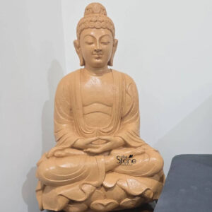 A two-foot-tall seated Buddha statue carved from warm-toned sandstone is depicted in a classic meditative posture facing the viewer directly. The statue exhibits a rich textured surface with visible natural grain and slight color variations inherent to the stone. Lord Buddha is seated on a detailed carved lotus pedestal symbolizing purity and spiritual awakening. His legs are crossed in the vajra position and his hands are resting in his lap in the dhyana mudra with palms facing upward and thumbs touching. The carving on the Buddha statue captures serene and benevolent facial features with eyes gently closed in contemplation and a slight compassionate smile. His head is adorned with traditional tightly curled ushnisha hair and elongated earlobes. The robe is draped simply over his left shoulder with subtle flowing lines carved to indicate the fabric folds. The statue is positioned against a plain light-colored wall in an indoor setting with a portion of a dark indistinct object visible on the right side of the frame. Natural light illuminates the statue from the front-left highlighting the intricate details and the tactile quality of the sandstone. This beautiful two-foot sandstone Buddha statue is an exemplary piece of traditional South Indian stonework perfectly suited to bring peace and tranquility to any space. The compact size makes it an excellent choice for a home shrine a personal meditation corner or a decorative focal point within a living area particularly for those seeking a high-quality Buddha murti in Kannur Kerala. The durable sandstone material and the profound expression of the sculpture make it a lasting symbol of mindfulness and spiritual harmony enhancing the ambiance of its surroundings with its serene presence.