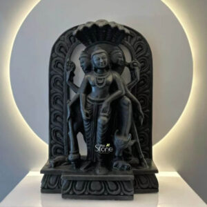 An eighteen-inch-tall finely crafted Blackstone Dattatreya statue stands as the central focus presented on a white elevated platform. The deity, representing the Hindu trimurti of Brahma, Vishnu, and Shiva combined is depicted in a standing posture facing forward with a serene benevolent expression. The statue is carved from deep black polished stone exhibiting a smooth texture and rich dark color that contrasts with the minimalist white background. Dattatreya has three faces each adorned with traditional ornaments and a crown symbolizing his omniscience. He is depicted with four arms holding symbolic attributes a mala and water pot in his upper hands and a danda and begging bowl in his lower hands representing his ascetic and divine nature. The Dattatreya statue is intricately detailed with flowing garments elaborate jewelry including multiple necklaces and armbands and a sacred thread visible across his chest. The carving captures the fine details of his hair and the ornamentation on all three crowns. The figure is positioned within a richly carved arched prabhavali that frames the deity and is supported by miniature pillars. The arch features a repeating scrollwork pattern and is topped by a stylized figure of a seated deity. At the base of the statue resting on the main pedestal are two small carved figures: a standing cow on the right and a seated figure on the left traditionally associated with Dattatreya iconography. The entire sculpture rests on a tiered base also adorned with a repeating carved pattern. This exquisite eighteen-inch Blackstone Dattatreya Statue is a perfect representation of divine knowledge and asceticism expertly crafted to serve as a revered murti for worship. The high-quality blackstone material and the profound level of detail in the carving, from the three faces to the delicate jewelry and attributes showcase the skill of the artisans. The compact eighteen-inch size makes it an ideal choice for home shrines personal altars or as a significant spiritual artifact within a temple setting particularly for devotees seeking a Dattatreya murti in Thane. The serene presence and traditional iconography of this statue make it a powerful focal point for spiritual practice and cultural appreciation.