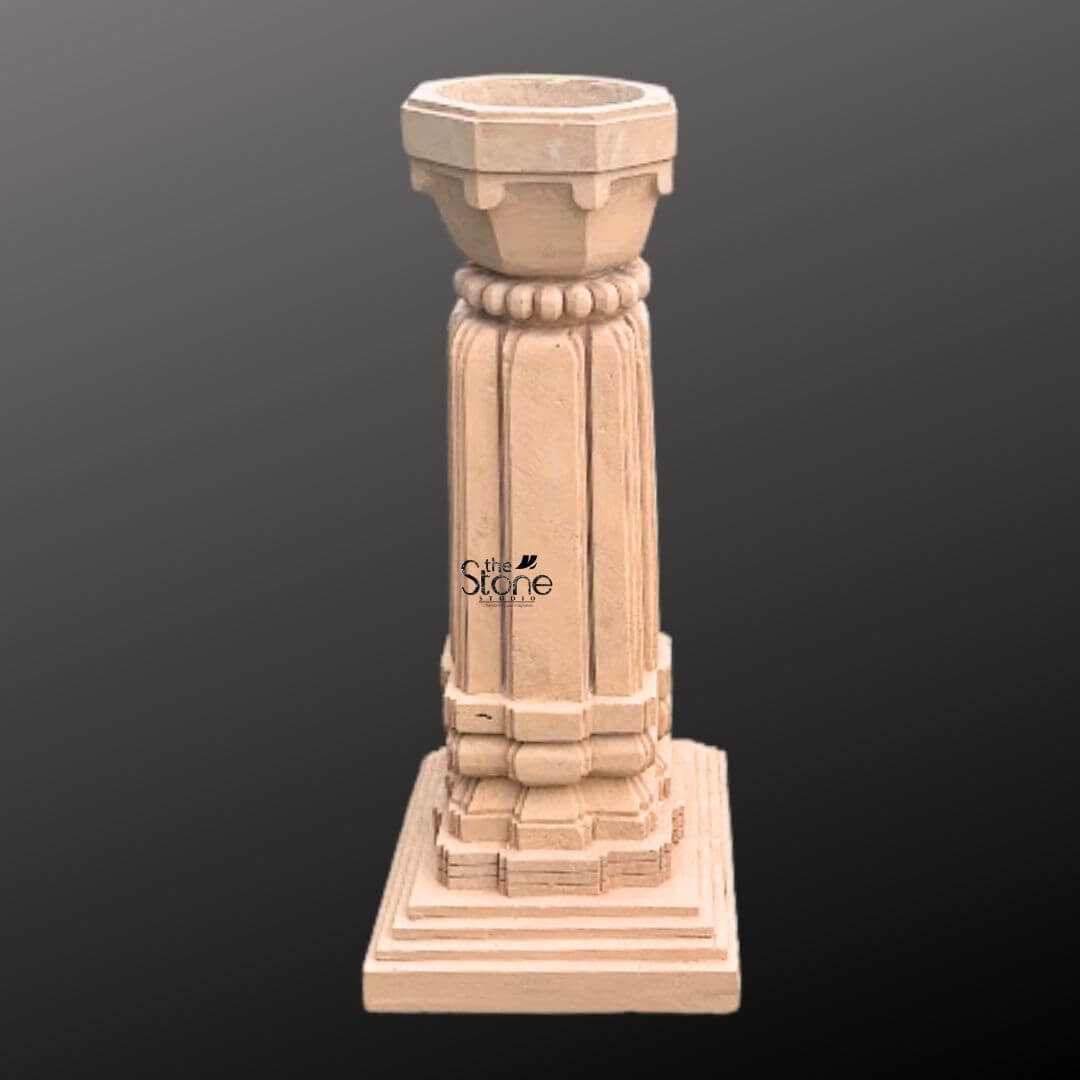 A three-quarter view photograph showcases a meticulously hand-carved Decorative Stone Tulsi Planter for the home standing approximately 3 feet tall. This traditional Indian planter is crafted from a single block of warm brown sandstone exhibiting a natural textured finish. The entire structure rests upon a substantial multi-tiered square base which provides excellent stability and a sense of grounded elegance making it a perfect addition to a home garden or courtyard. The main body of the planter features a beautifully proportioned square column adorned with classic vertical fluting on all four sides creating elegant lines that enhance its architectural presence. The sandstone surface displays subtle variations in color and grain highlighting the authentic artisanal quality of the stonework. The precision of the carving reflects a deep respect for traditional Indian temple architecture and design. Crowning the column is a beautifully shaped flared planter bowl designed to hold the sacred Tulsi plant and soil. This upper section transitions smoothly from the square column to a circular form featuring a decorative band of carved bead-and-reel molding just below the rim. The overall form is a harmonious blend of functionality and aesthetic grace making this sandstone Tulsi planter a revered and beautiful focal point for any home dedicated to traditional Indian culture and spirituality.