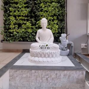 A beautifully composed photograph captures a serene indoor shrine featuring a Buddha and Fountain Fibre Statues standing as prominent examples of high-quality Fibre Statues in Bangalore. The central figure is a majestic 5-foot tall Buddha statue meticulously crafted from durable fibreglass with a smooth matte white finish that gives the appearance of carved marble. Lord Buddha is depicted in a traditional, peaceful seated posture known as vajrasana with both legs crossed and his right hand resting on his knee in the bhumisparsha mudra symbolizing his enlightenment. The Buddha statue is seated upon a substantial custom-built plinth that serves as the base for the entire installation. The plinth is faced with textured grey stacked stone tiles providing a rustic and grounded contrast to the smooth white fibreglass. The upper surface of the plinth is finished with polished white marble slabs creating a clean and elevated platform. To the right of the Buddha sits a matching multi-tiered cascade fountain also in a white stone finish, with water gently flowing down the bowls adding a soothing auditory element to the visual tranquility. The backdrop for this peaceful shrine is a stunning vertical garden wall completely covered in a dense tapestry of various green tropical plants. The lush foliage provides a rich, natural texture that complements the stone and fibreglass elements bringing an element of nature indoors which is a popular design trend in Bangalore homes. The entire setup is positioned on a light-colored stone floor emphasizing the reverence and care with which this Fibre Statues installation is presented within the residential space. The bright natural daylight from an unseen window highlights the intricate details of the carving and the freshness of the vertical garden.