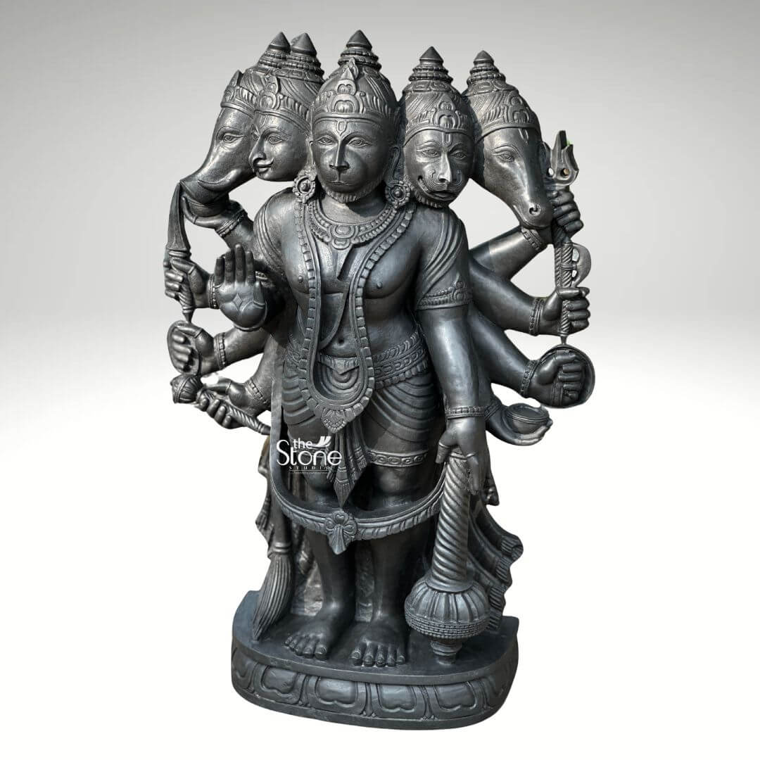 A three-quarter view studio photograph set against a plain light grey background displays a magnificent 3.5-foot Five Face Hanuman Stone Statue. This intricately hand-carved Vigraham is made from a single block of deep dark blackstone renowned for its durability and fine grain. The statue depicts the five-faced form of Lord Hanuman known as Panchamukhi Hanuman standing firmly on a multi-tiered ornate circular base. The central face is that of Hanuman himself looking forward with a serene protective expression adorned with traditional jewelry and a crown. Radiating outwards from the central head are four other divine faces: to the east the boar face to the south the lion face to the west the eagle face and to the north the horse face. The statue is multi-armed totaling ten arms that hold various divine attributes each rendered with exquisite detail. The main central right hand is raised in the Abhaya mudra while the main central left hand holds a mace that rests on the base. The other eight hands hold a conch discus sword shield noose hook a mountain and a vessel. The deity is adorned with elaborate traditional South Indian carvings of necklaces armlets wristlets a sacred thread and a detailed dhoti all chiseled with precision into the dark stone. The fine texture of the blackstone enhances the depth and shadow of the carvings highlighting the complex iconography of this formidable yet benevolent form of Hanuman. This Five Face Hanuman Stone Statue is an exceptional example of traditional Indian temple sculpture suitable for sanctum placement or as a grand focal point in a home shrine or spiritual space. The 3.5-foot height makes it a commanding presence embodying the combined power of the five deities represented by the faces. The blackstone material gives the statue a timeless ancient feel ensuring it will be a cherished piece for generations. The statue's detailed iconography from the expressions on the five faces to the specific items held in the ten hands makes it a significant subject for study and devotion. The expert craftsmanship transforms the raw stone into a vibrant sacred form.