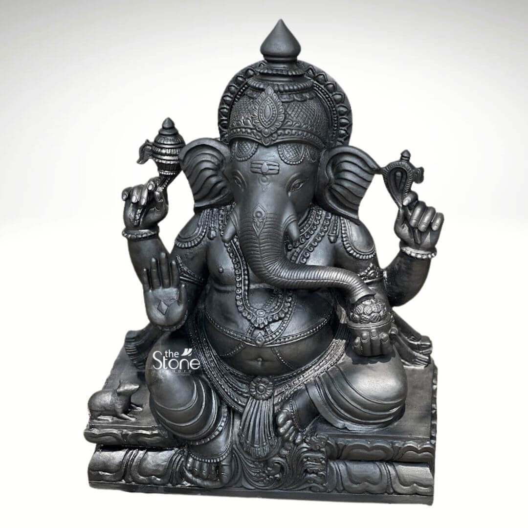 A magnificent 2.5-foot-tall Ganapathi black stone vigraham stands as a profound example of traditional South Indian sculptural art perfectly scaled for a home shrine or temple sanctum. Hand-carved from a single block of deep blackstone the statue depicts the Hindu deity Ganesha in a traditional seated posture known as the Lalitasana pose, with his right leg folded and left leg slightly raised. The dark polished stone surface exhibits a subtle cool sheen under the studio lighting highlighting the smooth texture of the material. The artisan has lavished attention on the intricate details from the ornate crown and multiple necklaces adorning his form to the delicate folds of his flowing dhoti and the individual petals of the lotus throne he occupies. Ganesha is depicted with four arms each holding a symbolic item with great precision his upper right hand holds an ankusha his upper left holds a pasha and his lower right hand is in the Abhaya Mudra bestowing blessings upon devotees. His lower left hand holds a modak resting in his lap with his trunk gently curled to sample the offering. The statue features a prominent finely carved prabhavali behind the deity adorned with traditional patterns and culminating in a kirtimukha at the apex. His loyal vahana the mouse is seated attentively at his feet to the left completing the divine composition. This black stone Ganapathi vigraham strikes an ideal balance being large enough to serve as a central deity in a home puja room yet detailed enough to be appreciated as a significant decorative piece. The substantial weight and durable stone material ensure its longevity making it a lasting investment for spiritual practice or cultural decor. The clean studio photograph against a plain background emphasizes the statue's three-dimensional quality and the artisan's meticulous work on the serene facial expression and decorative elements, perfectly illustrating the grace and cultural richness that a well-crafted black stone Ganesha statue brings to any consecrated space.