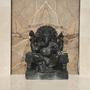 A three-quarter view photograph captures a magnificent Ganapathi Statue meticulously hand-carved from a single piece of deep dark basalt stone. Standing approximately 3 feet tall this imposing statue depicts Lord Ganesha in a traditional seated posture known as lalitasana with one leg folded and the other resting comfortably. The statue's smooth matte finish accentuates the natural rich color of the stone and the exceptional precision of the sculptural details. The deity is adorned with elaborate deeply-cut ornamentation across his form including multiple necklaces armbands wristlets and anklets all rendered in the durable black stone. He wears a richly carved crown and a sacred thread across his chest. He has four arms his upper right hand holds a parashu and his upper left holds a pasha. His lower right hand is raised in the abhaya mudra while his lower left hand cradles a modaka which his gracefully curved trunk is poised to taste. This Ganapathi statue is enshrined within a custom-built recessed niche on a textured exterior wall showcasing its suitability for a home shrine or garden setting in Dubai. The niche features a simple frame of polished cream-colored stone and a backdrop of textured beige marble with subtle white veining, which provides a beautiful contrast to the dark stone of the statue. The entire composition sits on a substantial polished marble shelf emphasizing its revered placement within a contemporary Dubai architectural context.