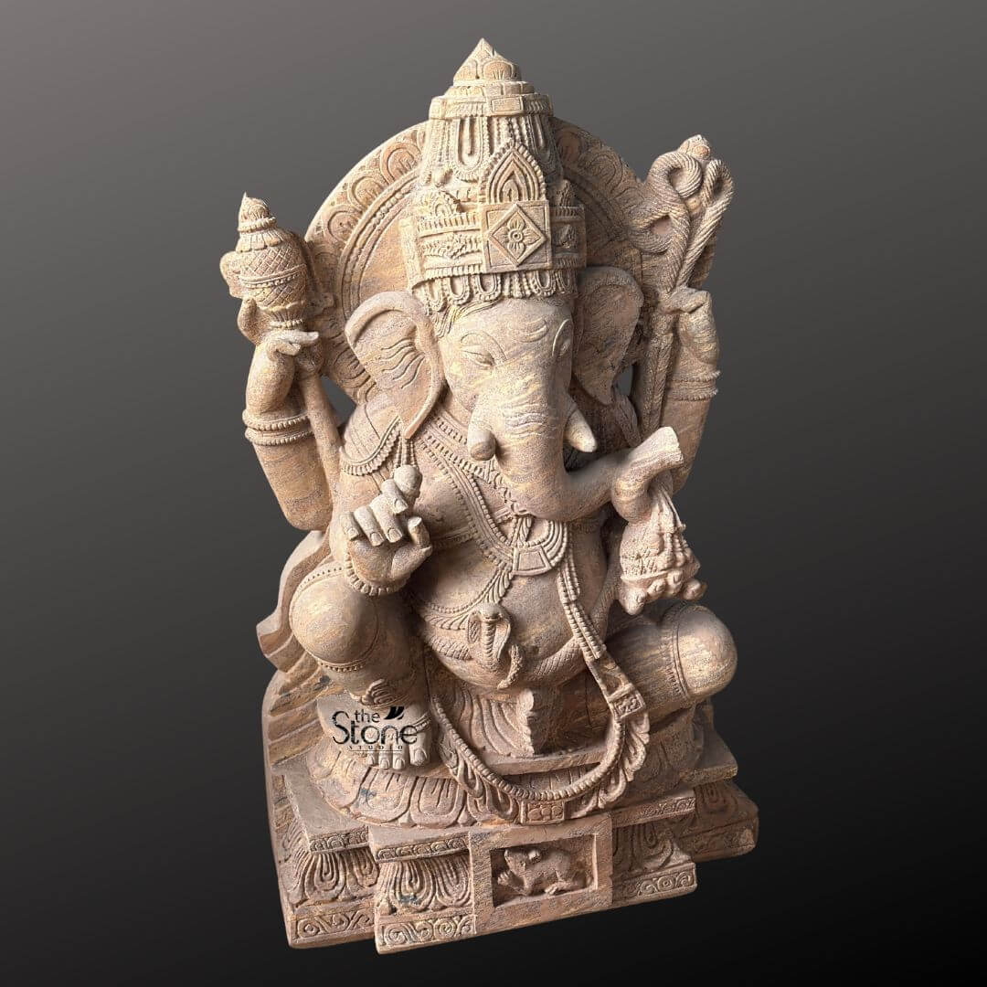 A detailed eye-level photograph of a magnificent Ganapathi Stone Vigraham meticulously hand-carved from a single block of warm brown sandstone. The statue stands approximately four feet tall and is depicted in a traditional seated posture known as lalitasana with one leg gracefully folded and the other resting on a decorated base. The four-armed deity is seated upon a carved multi-tiered lotus pedestal which is further embellished with intricate floral and geometric patterns and features a small detailed carving of Ganesha's vehicle the mouse centrally at the base. The sandstone exhibits a natural slightly porous texture and warm color variations typical of the material showcasing the artisan's skill in achieving fine detail. Ganapathi is richly adorned with abundant deeply carved traditional jewelry and ornaments all rendered in the same sandstone. He wears a detailed jeweled mukuta featuring a prominent central gem and peacock motifs multiple necklaces of varying lengths elaborate armlets wristlets and anklets. His sacred thread is intricately carved across his torso. The deity holds symbolic attributes in his four hands: his upper right hand holds an axe and his upper left hand holds a noose while his lower right hand is raised in a gesture of blessing and his lower left hand holds a sweet towards his trunk. The trunk itself curves elegantly to the left holding the modak. This commanding 4 feet statue embodies the spiritual essence and iconographic details of the remover of obstacles making it a suitable and sacred focal point for a home shrine or temple sanctum. The face of Ganapathi is serene and benevolent with downcast eyes conveying deep wisdom and compassion. The careful carving of every element from the tassels of his garments to the petals of the lotus throne reflects centuries-old sculpting traditions. The overall composition is balanced and reverent highlighting the divine form of Lord Ganesha. The photograph captures the statue against a plain dark grey studio background emphasizing the detailed craftsmanship and the natural beauty of the sandstone.