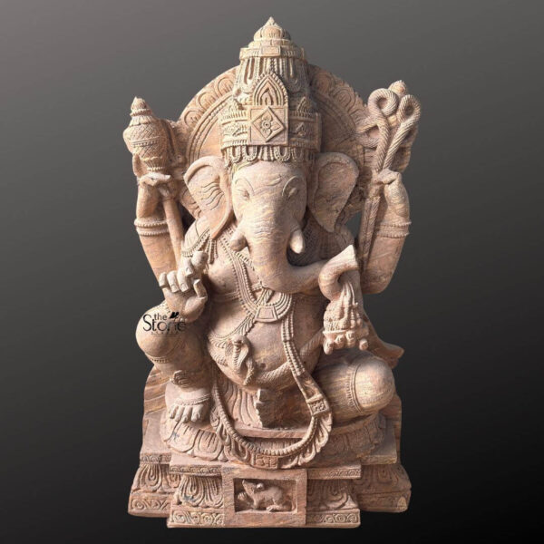 A full eye-level photograph of a magnificent Ganapathi Swamy Stone Vigraham meticulously hand-carved from a single block of warm beige sandstone. The statue stands approximately four feet tall and is depicted in a traditional seated posture known as lalitasana with one leg gracefully folded and the other resting on a decorated base. The four-armed deity is seated upon a carved multi-tiered lotus pedestal which is further embellished with intricate floral and geometric patterns and features a small detailed carving of Ganesha's vehicle the mouse centrally at the base. The sandstone exhibits a natural slightly porous texture and warm color variations typical of the material showcasing the artisan's skill in achieving fine detail. Ganapathi Swamy is richly adorned with abundant deeply carved traditional jewelry and ornaments all rendered in the same sandstone. He wears a detailed jeweled mukuta eaturing a prominent central gem and peacock motifs multiple necklaces of varying lengths elaborate armlets wristlets and anklets. His sacred thread is intricately carved across his torso. The deity holds symbolic attributes in his four hands his upper right hand holds an axe and his upper left hand holds a noose while his lower right hand is raised in a gesture of blessing and his lower left hand holds a sweet towards his trunk. The trunk itself curves elegantly to the left holding the modak. This commanding 4 feet statue embodies the spiritual essence and iconographic details of the remover of obstacles making it a suitable and sacred focal point for a temple sanctum. The face of Ganapathi Swamy is serene and benevolent with downcast eyes conveying deep wisdom and compassion. The careful carving of every element from the tassels of his garments to the petals of the lotus throne reflects centuries-old sculpting traditions. The overall composition is balanced and reverent highlighting the divine form of Lord Ganesha. The photograph captures the statue against a plain dark grey studio background emphasizing the detailed craftsmanship and the natural beauty of the sandstone.