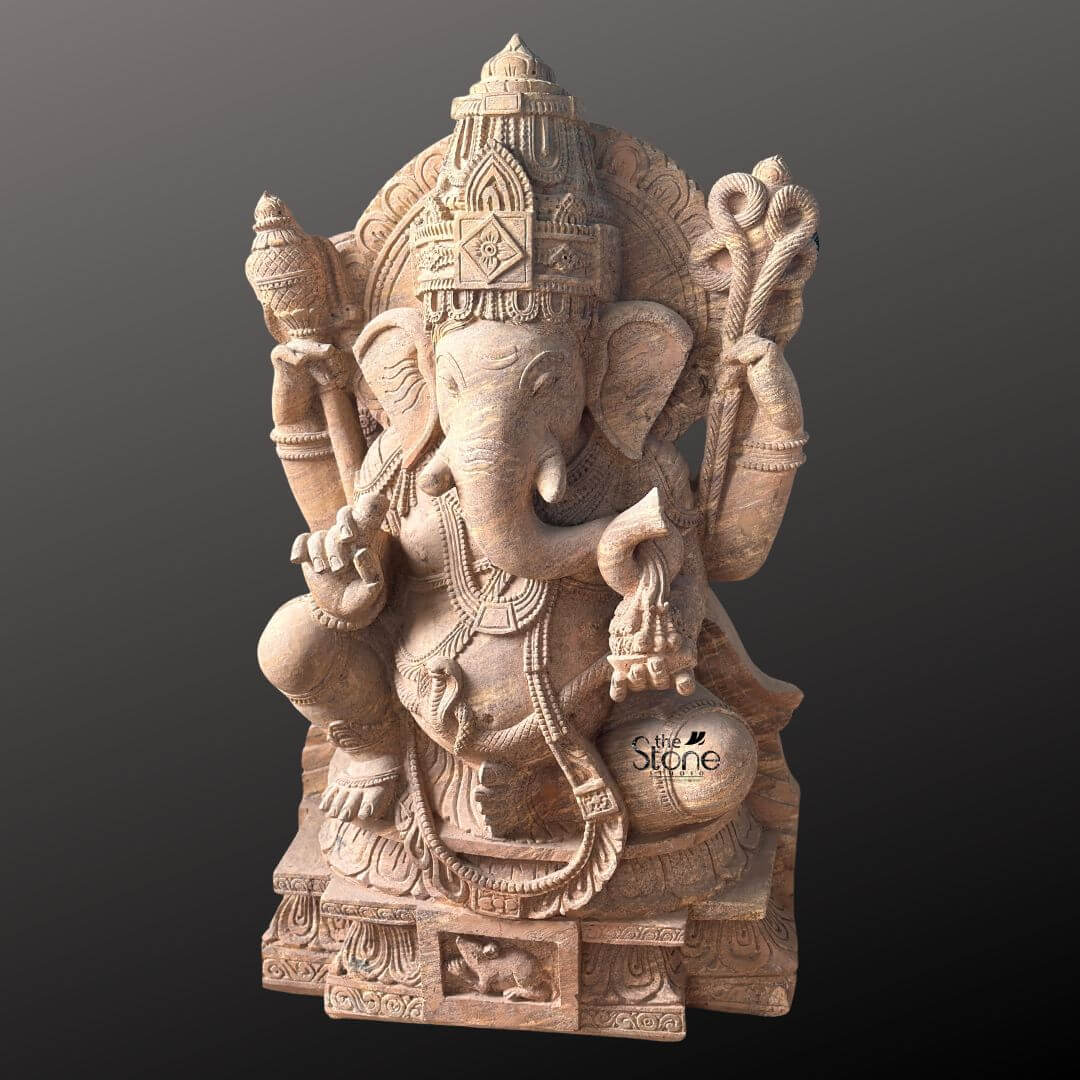 A full eye-level photograph of a magnificent Ganapathi Swamy Stone Vigraham meticulously hand-carved from a single block of warm brown sandstone. The statue stands approximately four feet tall and is depicted in a traditional seated posture known as lalitasana with one leg gracefully folded and the other resting on a decorated base. The four-armed deity is seated upon a carved multi-tiered lotus pedestal which is further embellished with intricate floral and geometric patterns and features a small detailed carving of Ganesha's vehicle the mouse centrally at the base. The sandstone exhibits a natural slightly porous texture and warm color variations typical of the material showcasing the artisan's skill in achieving fine detail. Ganapathi Swamy is richly adorned with abundant deeply carved traditional jewelry and ornaments all rendered in the same sandstone. He wears a detailed jeweled mukuta featuring a prominent central gem and peacock motifs multiple necklaces of varying lengths elaborate armlets wristlets and anklets. His sacred thread is intricately carved across his torso. The deity holds symbolic attributes in his four hands: his upper right hand holds an axe and his upper left hand holds a noose while his lower right hand is raised in a gesture of blessing and his lower left hand holds a sweet towards his trunk. The trunk itself curves elegantly to the left holding the modak. This commanding 4 feet statue embodies the spiritual essence and iconographic details of the remover of obstacles making it a suitable and sacred focal point for a home shrine or temple sanctum. The face of Ganapathi Swamy is serene and benevolent with downcast eyes conveying deep wisdom and compassion. The careful carving of every element from the tassels of his garments to the petals of the lotus throne reflects centuries-old sculpting traditions. The overall composition is balanced and reverent highlighting the divine form of Lord Ganesha. The photograph captures the statue against a plain dark grey studio background emphasizing the detailed craftsmanship and the natural beauty of the sandstone.