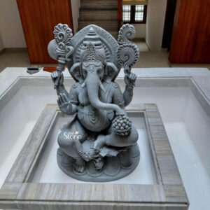 A full-view photograph captures a magnificent 4-foot tall Ganesha Fibre Statue meticulously crafted and positioned as a central home decor element in a modern residence in Thiruvananthapuram. The statue is made from high-quality fibreglass possessing a cool grey stone-like finish that exudes a sense of ancient elegance while remaining lightweight and durable. Lord Ganesha is depicted in a traditional seated posture known as lalitasana with one leg folded and the other resting comfortably exuding divine grace and solemnity. The deity is adorned with elaborate deeply cut ornamentation across his four-armed form. He wears a richly decorated crown multiple strands of intricate necklaces armlets wristlets and anklets all rendered with exceptional precision in the grey stone-finish fibreglass. His upper right hand holds a parashu and his upper left holds a pasha. His lower right hand is raised in the abhaya mudra while his lower left hand cradles a modaka which his gracefully curved trunk is poised to taste. This Ganesha Fibre Statue is seated upon an ornate multi-tiered lotus pedestal providing a stable and decorative base. The entire setup is enshrined within a custom-built recessed marble alcove in a contemporary hallway emphasizing its revered placement. The background shows a warm wooden door on the left a light-filled window with modern blinds and a staircase with marble steps leading upward creating a harmonious blend of traditional spirituality and modern interior design. The craftsmanship highlights the spiritual significance and artistic beauty of this large-scale home decor piece.