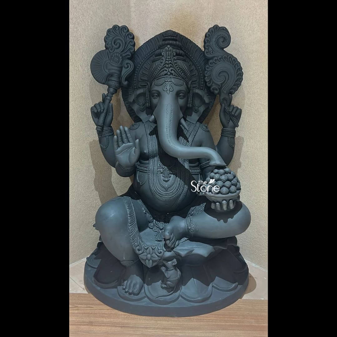 A magnificent four-foot-tall Ganesha Fibre Statue stands as a divine centerpiece showcasing exceptional artistry and craftsmanship. The statue is rendered in a deep matte charcoal-grey finish mimicking the appearance of polished black stone while retaining the lightweight and durable properties of fibreglass. Ganesha is depicted in a traditional seated posture known as the Lalitasana pose with his right leg gracefully folded and his left leg slightly raised resting on a sturdy integrated lotus-petal-carved pedestal. The artisan has lavished attention on the intricate details from the elaborate crown and multiple necklaces adorning his form to the delicate folds of his flowing dhoti all highlighted by the statue’s uniform dark tone and subtle highlights from the focused indoor lighting. Ganesha is depicted with four arms, each holding a symbolic item: his upper right hand holds an ankusha his upper left holds a pasha and his lower right hand is in the Abhaya Mudra bestowing blessings. His lower left hand holds a modak resting in his lap. Behind the deity a large finely carved prabhavali is adorned with traditional patterns and culminates in a kirtimukha at the apex adding grandeur and spiritual significance to the composition. The statue’s expression is one of serene benevolence with large, defined eyes and a gently curved trunk holding the modak bowl. This four-foot Ganesha Fibre Statue is an ideal size for a prominent home shrine a temple hall or a garden sanctuary in Trichy where it would serve as a powerful focal point for devotion and cultural pride. The substantial height commands attention while the durable fibreglass material ensures its longevity making it a practical and revered choice for placement in various environments. The photograph against a plain textured wall emphasizes the statue's three-dimensional quality and the artisan's meticulous work on the decorative elements perfectly illustrating the grace and cultural richness that a well-crafted Ganesha statue brings to any space.
