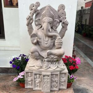 A full eye-level photograph of a magnificent Ganesha Stone Statue standing approximately four feet tall positioned outdoors against a plain white wall. The statue is meticulously hand-carved from a single block of off-white sandstone exhibiting a natural matte texture that highlights the intricate details of the craftsmanship. Ganesha is depicted in a traditional seated posture known as lalitasana with one leg folded and the other slightly pendant resting upon a carved lotus pedestal. He is adorned with extensive finely detailed traditional jewelry including multiple necklaces armlets wristlets anklets and an elaborate jeweled mukuta that sits atop his head all sculpted in high relief from the sandstone. The four-armed deity holds symbolic attributes in his hands an axe and a lotus in his upper right and left hands respectively while his lower right hand is raised in a gesture of blessing and his lower left hand holds a sweet towards his trunk. The face of Ganesha is serene and composed with downcast eyes conveying a sense of deep wisdom and divinity. His form is sculpted with careful attention to anatomical detail from the gentle contours of his body to the graceful curve of his trunk which rests upon the modak. The entire sculpture reflects the rich tradition of Indian temple art capturing the spiritual essence and iconographic details of the remover of obstacles. The statue is mounted on a rectangular sandstone plinth base which is decorated with carved panels depicting small standing figures and other motifs in relief. The setting appears to be the entrance area of a home or a garden in Noida with potted plants featuring white purple and pink flowers placed at the base of the statue on both sides adding a touch of natural color. The ground in the foreground is paved with reddish-brown stone tiles. The natural daylight illuminates the statue casting soft shadows that emphasize the depth and beauty of the carvings. This four-feet Ganesha statue in off-white sandstone is a fine example of traditional Indian stone carving suitable as a prominent spiritual and decorative piece for an outdoor shrine or a home entrance.