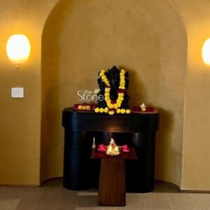 A serene warm-lit shrine in Rajkot centers on a beautifully crafted 18-inch Sitting Ganesha Stone Statue. The statue carved from a smooth dark blackstone that is characteristic of South Indian sculptural traditions depicts the Hindu deity Ganesha in a seated, meditative postur. His four arms hold traditional attributes an ankusha and pasha in the upper hands, a modak in one lower hand and the other lower hand in Abhaya mudra. The statue exhibits intricate detail, with fine carving on the jewelry crown and the texture of the dhoti all highlighted by the soft ambient light of the alcove. The Ganesha statue rests upon a polished dark wood console table within a recessed arched alcove with textured mustard-yellow plastered walls. The shrine area is richly decorated for worship. The statue is adorned with a prominent long garland of fresh yellow and white marigold flowers that drapes around his neck and falls to his lap. In front of the deity on a small separate dark wood offering stand a traditional brass oil lamp with multiple wicks is lit casting a flickering golden glow. Beside the lamp are small offerings of kumkum haldi loose marigold petals and a few small fruits like bananas presented on a decorative brass plate. The overall atmosphere of the shrine is one of deep peace and spiritual devotion making it a fitting focal point within a traditional Indian home in Rajkot. The dark stone of the Ganesha statue provides a powerful contrast against the warm textured wall while the fresh yellow marigolds add a vibrant touch of color and life. The arrangement demonstrates a classic home puja setup showcasing the 18-inch blackstone statue as a central object of worship. The lighting from a nearby warm-toned wall sconce emphasizes the smooth contours and detailed carvings of the deity creating a sacred space for daily prayer and reflection.
