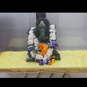 A front-facing photograph captures a beautifully adorned 15-inch Green Ganesha Idol for Home meticulously placed for worship in a sacred corner. The statue hand-carved from a single block of deep rich green stone depicts Lord Ganesha in a traditional seated posture. The cool natural tone and smooth texture of the stone are visible beneath the vibrant offerings highlighting the fine craftsmanship of the murti. The deity is lovingly decorated with fresh flower garlands that create a striking contrast against the dark green stone. A prominent garland of white fragrant flowers drapes elegantly around Ganesha's neck and shoulders covering his torso. Interwoven with this white garland are bright clusters of vivid orange marigold flowers adding a warm traditional touch. A few stems of deep purple orchids are carefully placed near his right hand introducing an element of exotic grace to the arrangement. The divine form of the remover of obstacles is reverently presented with his large expressive ears the graceful curve of his trunk and the traditional tilak mark on his forehead clearly discernible. The statue sits on a simple dark green plinth which in turn rests upon a small table covered in a shimmering yellow cloth with gold flecks scattered with loose marigold petals and purple orchid blooms. The simple background ensures the focus remains entirely on the beautifully decorated Green Ganesha Idol making it a perfect centerpiece for a home shrine.