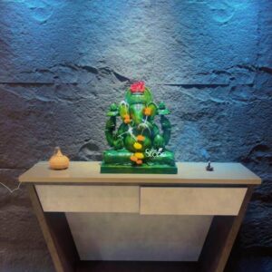 A straight-on medium shot photograph capturing a meticulously hand-carved Green Ganesha Statue standing approximately 21 inches tall positioned centrally on a modern two-tone console table. The statue is carved from a rich deep green stone showcasing a polished finish that highlights the natural variations and texture of the material. Ganesha is depicted in a traditional seated posture lalitasana with one leg folded and the other slightly pendant. His trunk curves gracefully to the left holding a modak and his four arms hold symbolic attributes including an axe a lotus and a pasha. The statue rests on a simple integrated plinth of the same green stone. The Ganesha statue is adorned with a fresh garland of small bright orange and yellow marigold flowers contrasting beautifully with the dark green stone and draped around his neck and chest. A single vibrant red hibiscus flower is placed atop his ornate crown. The statue is centered on the console table which features a warm dark wood frame and two drawers with a light grey textured front. The console has a minimalist trapezoidal base and sits on a light-colored tiled floor. The background provides a dramatic setting for the green deity. It is a textured accent wall composed of rough irregular dark grey stone blocks. The entire wall is dramatically illuminated from above by recessed LED cove lighting casting a cool deep blue wash over the rugged stone texture which makes the green Ganesha statue stand out as the clear focal point. To the left of the statue on the console table is a small rounded beige aroma diffuser, and to the right is a small dark incense burner with a wisp of smoke rising. The scene conveys a sense of modern spirituality and reverence making it an ideal setup for a home shrine.