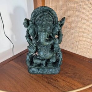 A medium shot captures a beautifully crafted 9-inch Green Stone Ganesha Statue positioned as a focal point on a polished dark wooden table. The statue is hand-carved from a deep rich greenstone possibly a type of soapstone known for its smooth slightly mottled texture and cool dark hue. Ganesha the elephant-headed Hindu deity is depicted in a seated meditative posture resting on a multi-tiered ornate circular lotus base. The statue exhibits intricate detail with elaborate carvings covering his crown multiple necklaces, armlets, wristlets, and the flowing patterns of his dhoti all chiseled with precision into the vibrant green stone. His trunk is gracefully curved to the left, holding a sweetmeat. Ganesha is multi-armed totaling four arms each holding a traditional attribute. His upper right hand holds a goad and his upper left hand holds a noose. His lower right hand is raised in the Abhaya mudra and his lower left hand holds a bowl of sweets. The dark deep green of the stone provides a striking contrast against the warm, reddish-brown grain of the wooden table making the statue stand out prominently. The natural light from a nearby source highlights the smooth polish of the stone and the depth of the carvings emphasizing the divine form and the artisan's skill. This 9-inch Green Stone Ganesha Statue exemplifies the high quality of traditional stone carving available in Bangalore a city renowned for its rich cultural heritage and craftsmanship. The specific type of greenstone used gives the murti a unique aesthetic blending the spiritual significance of the deity with the natural beauty of the stone. The statue's size makes it an ideal choice for a home puja altar a living room display or as a thoughtful culturally-resonant gift. Its presence conveys a sense of peace auspiciousness and artistic tradition, making it a perfect representative of the stone art for which Bangalore is famous. The intricate details from the gentle smile on Ganesha's face to the patterns on his ornaments showcase the timeless appeal of this sacred art form.