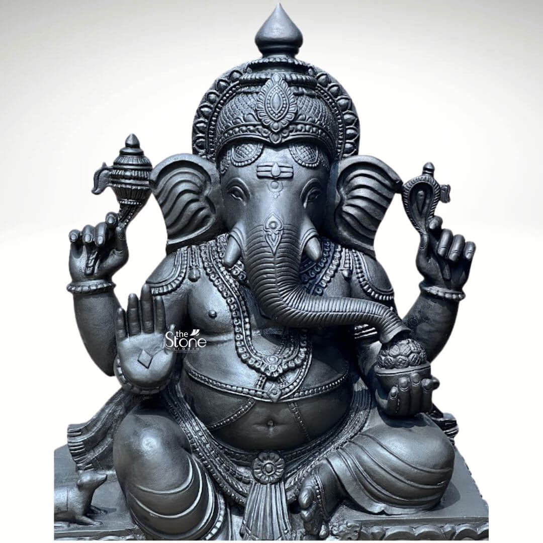 A magnificent hand-carved Ganesha statue standing 2.5 feet tall is presented against a clean neutral white studio background showcasing the sculpture in its entirety. The statue is carved from a single block of deep blackstone exhibiting a dark smooth and slightly cool-toned polished surface that accentuates the intricate details of the artisan's work. Ganesha is depicted in a traditional seated posture known as the Lalitasana pose with his right leg gracefully folded and left leg slightly raised resting on an integrated lotus-petal-carved pedestal. The artisan has lavished attention on the adornments from the elaborate crown and multiple necklaces to the armbands bracelets and the delicate folds of his flowing dhoti all meticulously detailed in the dark stone. Ganesha is depicted with four arms each holding a symbolic item with great precision. His upper right hand holds an ankusha his upper left holds a pasha and his lower right hand is in the Abhaya Mudra bestowing blessings upon devotees. His lower left hand holds a modak resting in his lap, with his trunk gently curved to sample the offering. The statue features a prominent finely carved prabhavali behind the deity, adorned with traditional patterns and culminating in a kirtimukha at the apex. His loyal vahana the mouse is seated attentively at his feet to the left completing the divine composition. This 2.5-foot hand-carved black stone Ganesha statue strikes an ideal balance being large enough to serve as a central deity in a home shrine or a small temple sanctum yet detailed enough to be appreciated as a significant decorative piece. The substantial weight and durable stone material ensure its longevity making it a lasting investment for spiritual practice or cultural decor. The clean studio photograph emphasizes the statue's three-dimensional quality and the artisan's meticulous work on the serene facial expression and decorative elements perfectly illustrating the grace and cultural richness that a well-crafted black stone Ganesha statue brings to any consecrated space.