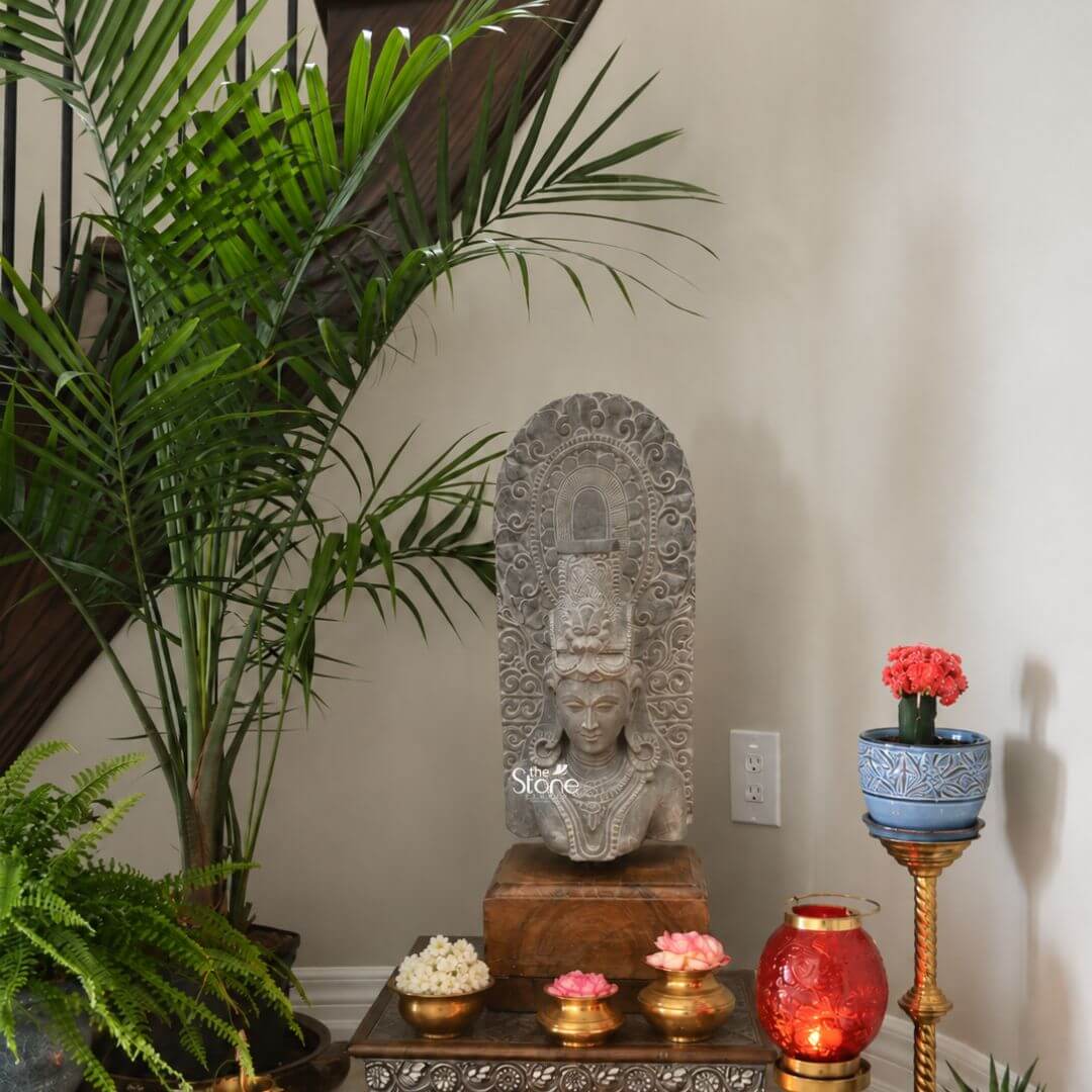 A magnificent thirty-inch hand-carved Lord Shiva bust in black stone stands as the revered centerpiece of a home shrine presented on a dark wooden pedestal. The sculpture meticulously chiseled from a single block of dark polished black stone exhibits a rich aged texture with natural variations and a weathered patina that underscores its antiquity. Lord Shiva is portrayed with a serene contemplative facial expression with eyes gently closed in meditation arched eyebrows and a subtle compassionate smile. His hair is intricately styled in a traditional jata forming a high crown adorned with delicate carvings of a crescent moon a serpent, and a central floral motif with further details of coiled locks falling onto his shoulders. The bust features elaborate ornamentation including large circular earrings and multiple layered necklaces with intricate pendants that rest upon his chest all hand-carved with precision. A sacred thread is visible across his torso. Behind the deity's head, a richly carved ornate prabhavali rises featuring detailed symmetrical floral and scrollwork patterns that frame his divine form. The craftsmanship showcases the traditional South Indian temple art style with deep undercuts and high relief that create a profound sense of depth and reverence. The entire sculpture rests securely on a dark polished wooden plinth providing a stable base. This exquisite thirty-inch hand-carved Lord Shiva black stone sculpture is a perfect testament to the timeless devotion and artistic skill of traditional Indian artisans. The large scale of the bust, combined with the deep black stone and the profound meditative expression makes it an awe-inspiring focal point for any spiritual space or home mandir. The detailed iconograph from the elaborate headdress to the intricate jewelry and the ornate prabhavali ensures this antique-style murti meets the highest aesthetic and spiritual requirements for those seeking a significant and beautiful representation of Lord Shiva. The natural color of the stone and the profound depth of the carving convey a sense of sanctity and spiritual presence.