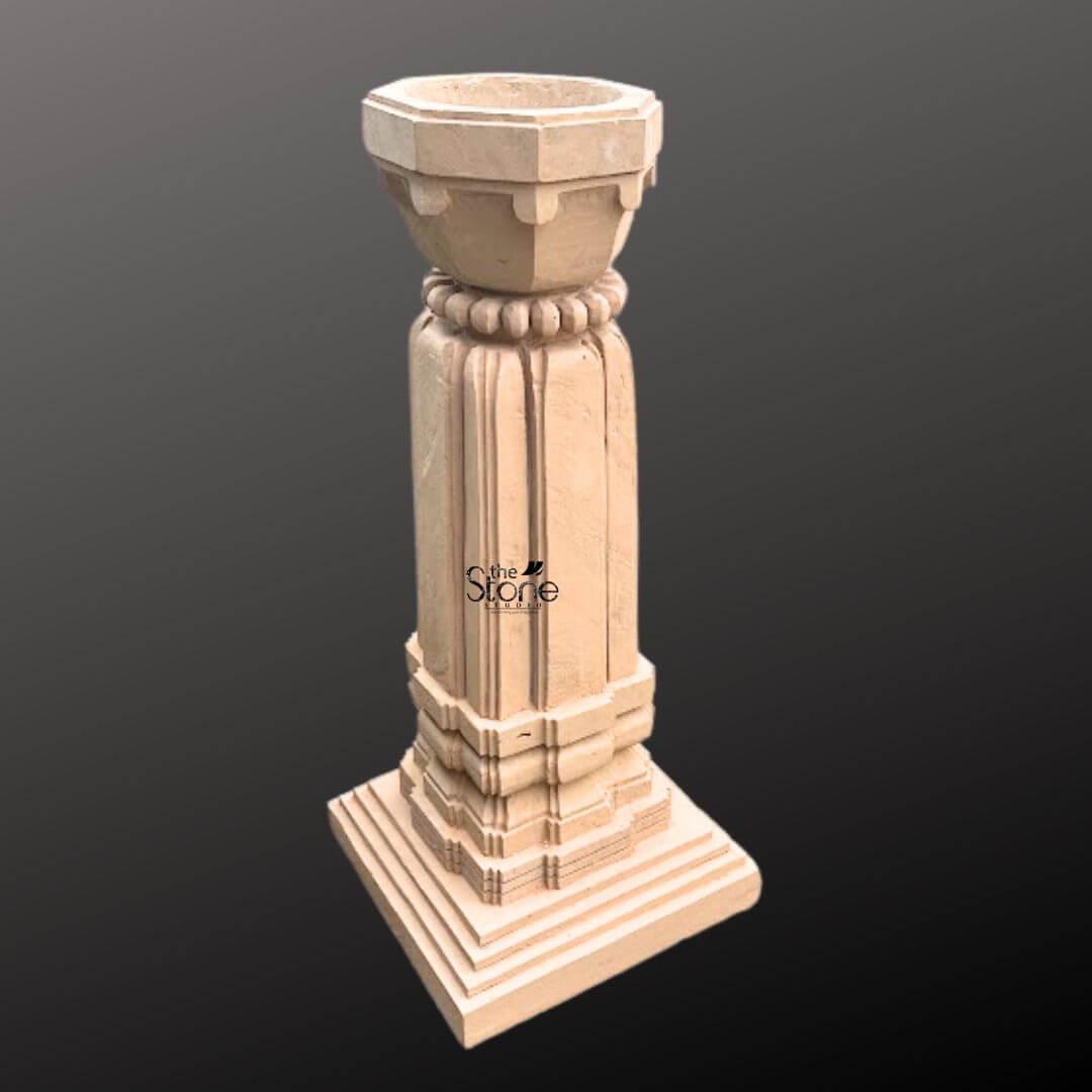 A three-quarter view photograph showcases a magnificent Hand Carved Stone Tulsi Planter standing approximately 3 feet tall against a simple dark gradient background. This traditional Indian planter is meticulously crafted from a single block of warm sandstone exhibiting a natural textured finish. The entire structure rests upon a substantial stepped square base providing stability and a sense of grounded grandeur perfect for a garden or courtyard setting. The main body of the planter features a beautifully proportioned square column adorned with classic vertical fluting on all four faces creating elegant lines that enhance its architectural presence. The sandstone surface displays subtle variations in color and grain highlighting the authentic artisanal quality of the hand-carving process. The precision of the work reflects a deep respect for traditional Indian stone carving techniques. Crowning the column is a beautifully shaped flared planter bowl designed to hold the sacred Tulsi plant and soil. This upper section transitions smoothly from the square column to a circular form featuring a decorative band of carved bead-and-reel molding just below the rim. The overall form is a harmonious blend of functionality and aesthetic grace making this sandstone Tulsi planter a revered and beautiful focal point for any home or garden.