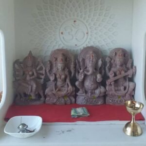 A neatly arranged home mandir or prayer space is presented featuring a set of four intricately carved pink stone god statues as its central focus. The statues rest upon a simple white shelf which is covered by a plain red cloth. Behind the deities the white wall is adorned with a subtle textured mandala pattern and a circular Om symbol serves as a spiritual backdrop. The four statues depicting Hindu deities are sculpted from a uniform warm pink-hued stone showcasing detailed craftsmanship in their traditional attire ornaments and serene facial expressions. From left to right the statues represent Durga Lakshmi Ganesha and Saraswati creating a powerful and balanced pantheon for daily worship. The deities are arranged in a specific order that is common in many Hindu households. On the far left sits the multi-armed goddess Durga mounted on her lion holding various weapons. Next to her is the goddess of wealth Lakshmi in a seated posture with hands in a blessing mudra. In the center is the elephant-headed god Ganesha depicted in a seated position with one hand raised in blessing. On the far right is the goddess of knowledge Saraswati playing a veena. Each statue sits on its own carved base contributing to a cohesive and sacred arrangement suitable for a home shrine in Mumbai. The foreground of the mandir shelf contains essential puja items that add to the atmosphere of devotion. To the left a simple white ceramic bowl holds a small silver oil lamp with a wick. To the right a traditional brass oil lamp stands its flame casting a soft warm light on the pink stone figures. A small stack of Indian currency notes is placed directly in front of the Ganesha statue as an offering. This entire setup provides inspiration and a beautiful example of how to create a consecrated space with traditional stone statues in a modern urban home.