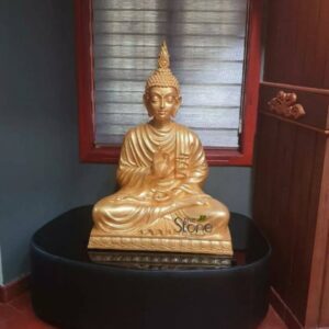 A three-foot-tall seated Buddha statue finished in a radiant metallic gold stands as the serene focal point of an indoor living space. The statue is positioned on a sleek black curved pedestal which rests on a terracotta-tiled floor. Lord Buddha is depicted in a traditional meditative posture with his legs crossed in the vajra position and his hands held in a gesture of teaching and exposition the vitarka mudra with the thumb and index finger of both hands forming a circle while the other fingers are extended upward. His face bears a calm benevolent expression with eyes gently closed in contemplation and a composed compassionate smile. The Buddha statue crafted from durable fibreglass exhibits a smooth polished golden surface that gleams under the ambient indoor light. His head is adorned with the traditional tightly curled ushnisha hair and elongated earlobes. The robe is draped simply over his left shoulder with subtle flowing lines forming the fabric folds and texture. The statue is positioned against a dark grey wall in the background which features a framed window with horizontal blinds partially visible behind the sculpture. To the right of the pedestal stands a dark wooden cabinet or pillar adding a touch of warm natural texture to the scene. This beautiful three-foot fibreglass Buddha statue is an exemplary piece of contemporary indoor decor perfectly suited to bring peace and tranquility to any home. The compact size makes it an excellent choice for a living room a personal meditation corner or a welcoming entryway particularly for those seeking a high-quality Buddha murti in Kannur Kerala. The durable fibreglass material ensures the sculpture's longevity and lightweight nature making it easy to place and maintain while the profound traditional iconography creates a lasting symbol of mindfulness and spiritual harmony within Kannur's modern living environments.