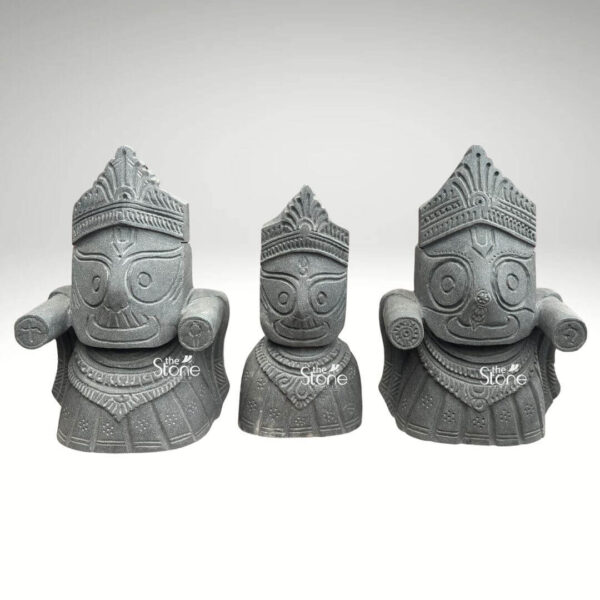 A three-quarter view studio photograph displays a masterfully hand-carved blackstone statue set of Lord Jagannath Lord Balabhadra and Devi Subhadra. The statues are sculpted from a single block of dark fine-grained blackstone exhibiting a smooth polished finish that highlights the deep rich color of the material. Each deity is depicted in the traditional iconic form associated with the Jagannath Temple of Puri characterized by large round staring eyes stylized facial features and distinctive tiered headgear. The statues are carved in a substantial blocky style conveying a sense of divine power and ancient tradition. The three figures are arranged in their traditional triad formation with Balabhadra and Jagannath flanking Subhadra. Lord Balabhadra and Lord Jagannath are depicted with their characteristic large square-cut heads and prominent, extended arms which are carved in a symmetrical outstretched manner. Devi Subhadra positioned centrally and slightly smaller, has a more rounded head and a serene smiling expression, with her arms represented in a simple folded posture. All three deities are seated upon a single integrated carved stone pedestal base providing a unified and stable composition. The intricate carving details of their traditional garments and ornate necklaces are rendered with precision contrasting beautifully with the smooth dark surface of the blackstone. The natural luster and deep hue of the blackstone material give the statue set a regal and spiritual presence making it a fitting centerpiece for a temple altar or a dedicated home shrine. The expert craftsmanship captures the spiritual essence and iconic forms of the deities showcasing the beauty of traditional Indian stone artistry. The smooth dark finish of the blackstone adds a sense of reverence and timelessness making this statue set a valuable addition to any collection of Hindu religious art.