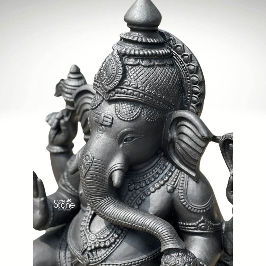 A close-up three-quarter view of a magnificent large Ganesha black stone idol standing 2.5 feet tall showcases the exceptional artistry and craftsmanship of traditional Indian stonework. The idol is carved from a single block of deep black soapstone exhibiting a dark smooth and subtly cool-toned polished surface that accentuates the intricate details of the artisan's work. Ganesha is depicted in a traditional seated posture known as the Lalitasana pose with his right leg gracefully folded and left leg slightly raised resting on an integrated lotus-petal-carved pedestal. The artisan has lavished attention on the adornments from the elaborate crown and multiple necklaces to the armbands bracelets and the delicate folds of his flowing dhoti all meticulously detailed in the dark stone. Ganesha is depicted with four arms each holding a symbolic item with great precision. His upper right hand holds an ankusha his upper left holds a pasha and his lower right hand is in the Abhaya Mudra bestowing blessings upon devotees. His lower left hand holds a modak resting in his lap with his trunk gently curled to sample the offering. The statue features a prominent finely carved prabhavali behind the deity adorned with traditional patterns and culminating in a kirtimukha at the apex adding grandeur and spiritual significance to the composition. The statue’s expression is one of serene benevolence with large defined eyes and a gentle, wise countenance. This 2.5-foot large black stone Ganesha idol strikes an ideal balance being large enough to serve as a central deity in a prominent home shrine or a small temple sanctum yet detailed enough to be appreciated as a significant decorative piece. The substantial weight and durable stone material ensure its longevity making it a lasting investment for spiritual practice or cultural decor. The clean studio photograph against a plain background emphasizes the statue's three-dimensional quality and the artisan's meticulous work on the serene facial expression and decorative elements perfectly illustrating the grace and cultural richness that a well-crafted black stone Ganesha statue brings to any space.