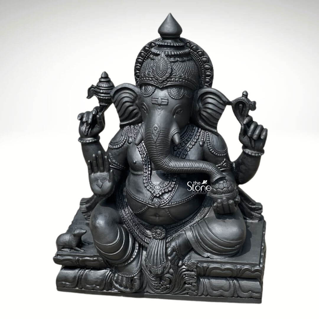 A magnificent 2.5-foot tall Lord Ganesh stone murti stands as a profound embodiment of traditional South Indian sculptural artistry perfectly scaled for a temple sanctum. Hand-carved from a single block of deep blackstone the statue depicts the Hindu deity in a traditional seated posture known as the Lalitasana pose with his right leg gracefully folded and left leg slightly raised. The dark polished stone surface exhibits a subtle cool sheen under the studio lighting highlighting the smooth texture of the material. The artisan has lavished attention on the intricate details from the ornate crown and multiple necklaces adorning his form to the delicate folds of his flowing dhoti and the individual petals of the lotus throne he occupies. Ganesha is depicted with four arms each holding a symbolic item with great precision. His upper right hand holds an ankusha his upper left holds a pasha and his lower right hand is in the Abhaya Mudra bestowing blessings upon devotees. His lower left hand holds a modak resting in his lap with his trunk gently curled to sample the offering. The statue features a prominent, finely carved prabhavali behind the deity adorned with traditional patterns and culminating in a kirtimukha at the apex. His loyal vahana the mouse is seated attentively at his feet to the left completing the divine composition. This black stone Ganesh murti strikes an ideal balance for temple use being substantial enough to command reverence in a smaller shrine yet detailed enough to be appreciated up close. The substantial weight and durable stone material ensure its longevity making it a lasting investment for spiritual practice and a timeless piece of temple decor. The clean studio photograph against a plain background emphasizes the statue's three-dimensional quality and the artisan's meticulous work on the serene facial expression and decorative elements perfectly illustrating the grace and cultural richness that a well-crafted black stone Ganesha brings to any consecrated space.