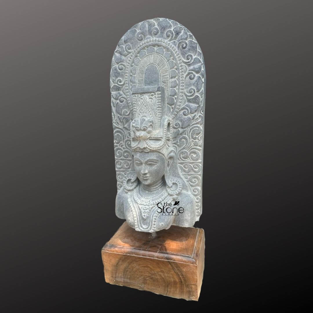 A thirty-inch hand-carved Lord Shiva head stone statue, presented as a detailed bust stands centrally on a dark wooden pedestal against a neutral grey-to-black gradient background. The sculpture is meticulously chiseled from a single block of deep black stone exhibiting a rich aged texture with natural variations and a weathered patina that underscores its antique character. Lord Shiva is portrayed with a serene contemplative facial expression characterized by eyes gently closed in meditation arched eyebrows and a subtle composed smile that conveys a profound sense of peace. The bust features elaborate ornamentation including large circular earrings and multiple layered necklaces with intricate pendants that rest upon his chest all hand-carved with precision. His hair is intricately styled in a traditional jata forming a high crown adorned with delicate carvings of a crescent moon a serpent and a central floral motif with further details of coiled locks falling onto his shoulders. Behind the deity's head, a richly carved, ornate prabhavali rises featuring detailed symmetrical floral and scrollwork patterns that frame his divine form. The craftsmanship showcases the traditional South Indian temple art style with deep undercuts and high relief that create a profound sense of depth and reverence. This magnificent thirty-inch hand-carved Lord Shiva head stone statue is a perfect testament to the timeless devotion and artistic skill of traditional Indian artisans. The large scale of the bust combined with the deep black stone and the profound meditative expression makes it an awe-inspiring focal point for any spiritual space or home mandir. The detailed iconography from the elaborate headdress to the intricate jewelry and the ornate prabhavali ensures this antique-style murti meets the highest aesthetic and spiritual requirements for those seeking a significant and beautiful representation of Lord Shiva. The natural color of the stone and the profound depth of the carving convey a sense of sanctity and spiritual presence.
