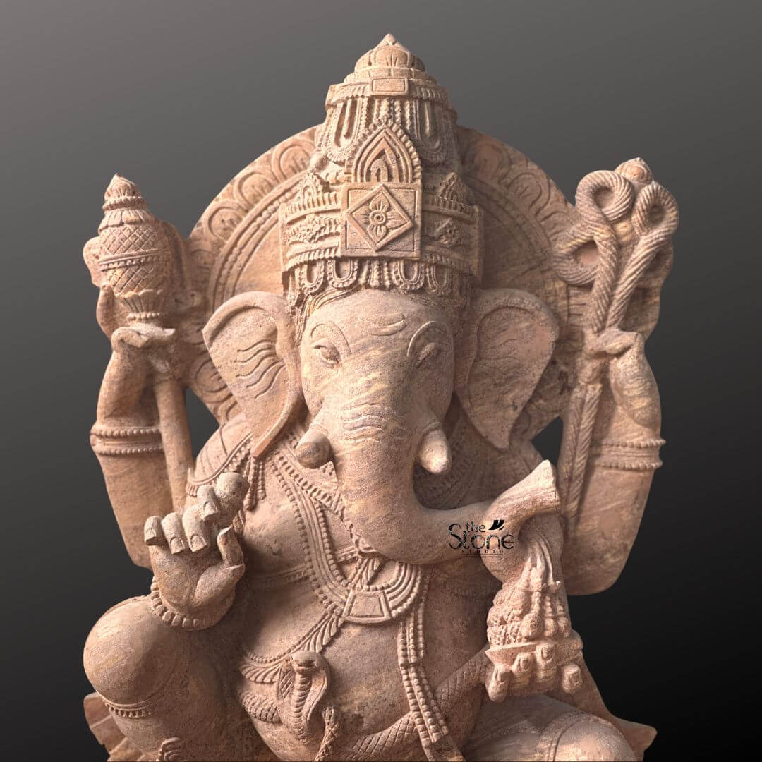 A detailed eye-level photograph of a magnificent Lord Vinayaka Stone Vigraham meticulously hand-carved from a single block of warm brown sandstone. The statue stands approximately four feet tall and is depicted in a traditional seated posture known as lalitasana with one leg gracefully folded and the other resting on a decorated base. The four-armed deity is seated upon a carved multi-tiered lotus pedestal which is further embellished with intricate floral and geometric patterns. The sandstone exhibits a natural slightly porous texture and warm color variations typical of the material showcasing the artisan's skill in achieving fine detail. Lord Vinayaka is richly adorned with abundant deeply carved traditional jewelry and ornaments all rendered in the same sandstone. He wears a detailed jeweled mukuta featuring a prominent central gem and peacock motifs multiple necklaces of varying lengths elaborate armlets wristlets and anklets. His sacred thread is intricately carved across his torso. The deity holds symbolic attributes in his four hands: his upper right hand holds an axe and his upper left hand holds a noose while his lower right hand is raised in a gesture of blessing and his lower left hand holds a sweet towards his trunk. The trunk itself curves elegantly to the left holding the modak. This commanding 4 feet statue embodies the spiritual essence and iconographic details of the remover of obstacles making it a suitable and sacred focal point for a home shrine or temple sanctum. The face of Lord Vinayaka is serene and benevolen with downcast eyes conveying deep wisdom and compassion. The careful carving of every element from the tassels of his garments to the petals of the lotus throne reflects centuries-old sculpting traditions. The overall composition is balanced and reverent highlighting the divine form of Lord Ganesha. The photograph captures the statue against a plain dark grey studio background emphasizing the detailed craftsmanship and the natural beauty of the sandstone.
