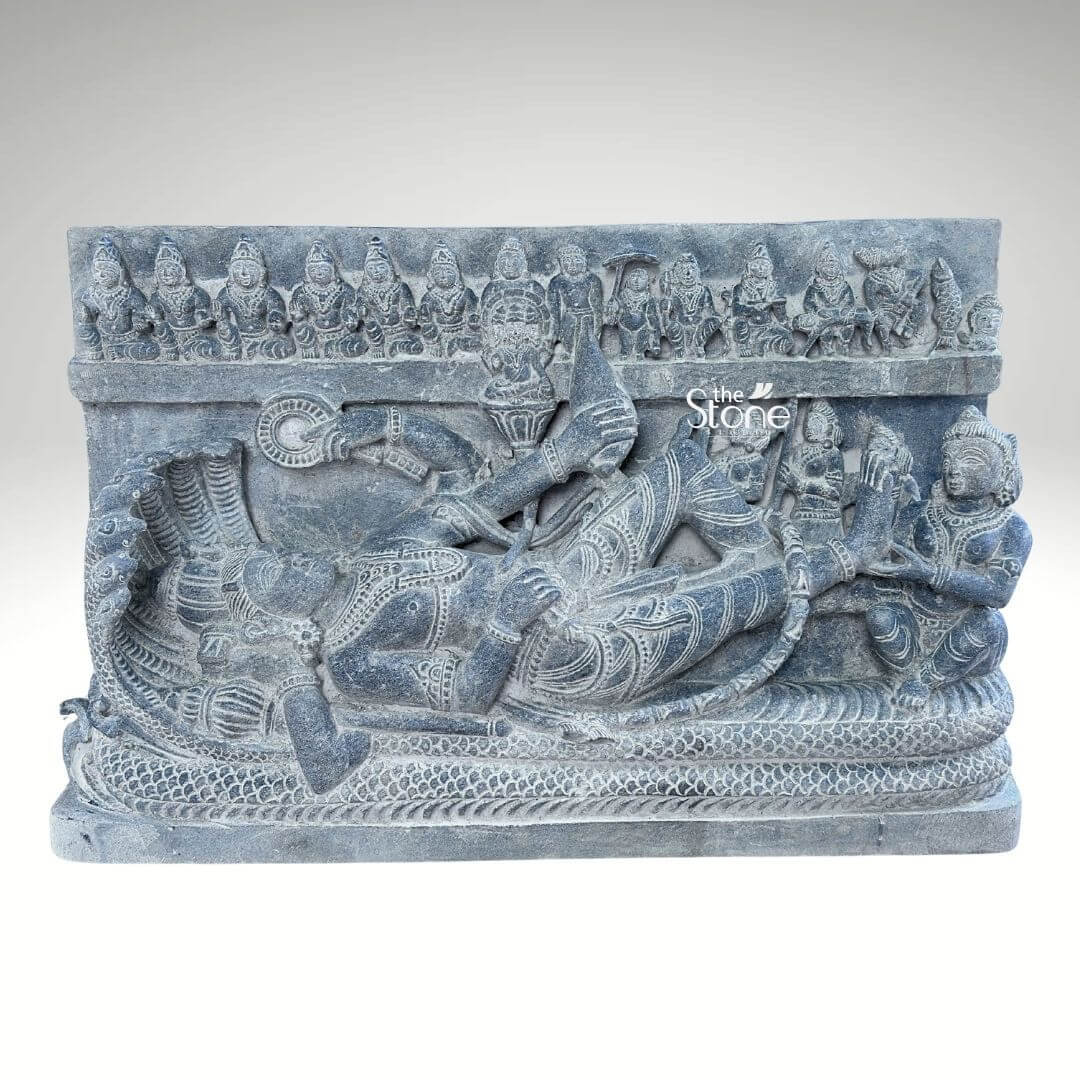 A detailed hand-carved blackstone statue approximately 30 inches tall depicting the Hindu deity Lord Vishnu in the Anantashayana posture reclining upon the cosmic serpent Sheshnag. The central figure of Vishnu lies peacefully on the coiled body of the multi-hooded serpent whose seven hooded heads fan out to form a protective canopy above the deity's crowned head. Vishnu is adorned with traditional jewelry a tall crown and elaborate garments with his right hand gently supporting his head while his left arm rests along his body holding a conch shell. The sculpture is carved in high relief showcasing the rich dark matte texture of the blackstone. Below the main reclining figure the coils of Sheshnag are intricately detailed forming a textured bed. Several attendant figures are carved into the stele including a seated Goddess Lakshmi massaging his foot at the base. Above the serpent canopy a frieze of miniature figures representing the ten avataras of Vishnu is carved horizontally across the top of the panel adding a comprehensive narrative to the divine iconography. The sculpture exhibits a refined finish with the natural variations in the stone's color and texture conveying a sense of ancient artistry and reverence consistent with a high-quality stone statue. The plain neutral grey background isolates the form and draws the viewer's eye directly to the complex narrative and divine iconography depicted in the stone. This detailed carving serves as an exemplary illustration for an article discussing Lord Vishnu reclining on Sheshnag stone statues.