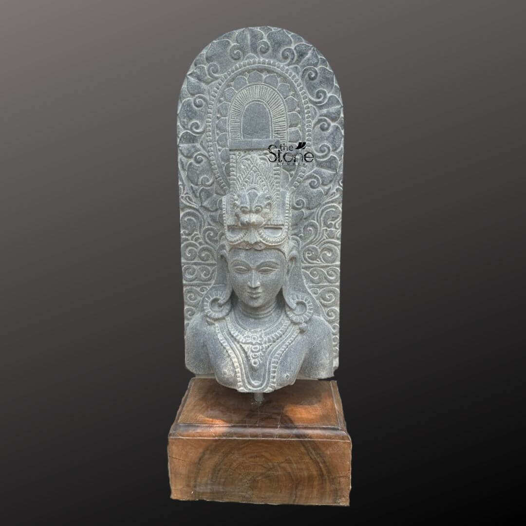 A magnificent thirty-inch hand-carved Lord Shiva bust or Mahadev stone bust statue stands as the revered centerpiece against a dark neutral gradient background. The sculpture meticulously chiseled from a single block of deep black stone exhibits a rich aged texture with natural variations and a weathered patina that underscores its antique character. Lord Shiva is portrayed with a serene, contemplative facial expression characterized by eyes gently closed in meditation arched eyebrows, and a subtle, composed smile that conveys a profound sense of peace and divine wisdom. The bust features elaborate ornamentation including large circular earrings and multiple layered necklaces with intricate pendants that rest upon his chest all hand-carved with precision. His hair is intricately styled in a traditional jata forming a high crown adorned with delicate carvings of a crescent moon a serpent and a central floral motif with further details of coiled locks falling onto his shoulders. Behind the deity's head a richly carved ornate prabhavali rises featuring detailed symmetrical floral and scrollwork patterns that frame his divine form. The craftsmanship showcases the traditional South Indian temple art style with deep undercuts and high relief that create a profound sense of depth and reverence. This exquisite thirty-inch hand-carved Mahadev black stone bust statue is a perfect testament to the timeless devotion and artistic skill of traditional Indian artisans. The large scale of the bust combined with the deep black stone and the profound meditative expression makes it an awe-inspiring focal point for any spiritual space or home mandir. The detailed iconography from the elaborate headdress to the intricate jewelry and the ornate prabhavali ensures this antique-style murti meets the highest aesthetic and spiritual requirements for those seeking a significant and beautiful representation of Lord Shiva. The natural color of the stone and the profound depth of the carving convey a sense of sanctity and spiritual presence making it a lasting addition to any sacred environment.