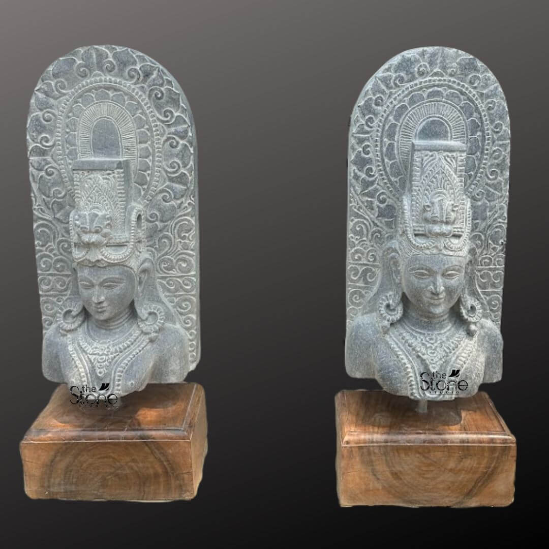 A diptych view of two identical thirty-inch hand-carved Lord Shiva bust statues or Mahadev stone statues is presented against a dark neutral gradient background. Each sculpture meticulously chiseled from a single block of black stone exhibits a rich aged texture with natural variations and a weathered patina that underscores its antique character. Lord Shiva is portrayed with a serene contemplative facial expression characterized by eyes gently closed in meditation arched eyebrows and a subtle composed smile that conveys a profound sense of peace and divine wisdom. The sculpture on the left shows the deity in a three-quarter profile while the one on the right presents a frontal view. The bust features elaborate ornamentation including large circular earrings and multiple layered necklaces with intricate pendants that rest upon his chest all hand-carved with precision. His hair is intricately styled in a traditional jata forming a high crown adorned with delicate carvings of a crescent moon a serpent and a central floral motif with further details of coiled locks falling onto his shoulders. Behind the deity's head a richly carved ornate prabhavali rises featuring detailed symmetrical floral and scrollwork patterns that frame his divine form. The craftsmanship showcases the traditional South Indian temple art style with deep undercuts and high relief that create a profound sense of depth and reverence. Each sculpture rests securely on a dark polished wooden plinth. This exquisite thirty-inch hand-carved Mahadev black stone statue is a perfect testament to the timeless devotion and artistic skill of traditional Indian artisans making it an ideal choice for both home and temple environments. The large scale of the bust combined with the deep black stone and the profound meditative expression makes it an awe-inspiring focal point for any spiritual space. The detailed iconography from the elaborate headdress to the intricate jewelry and the ornate prabhavali ensures this antique-style murti meets the highest aesthetic and spiritual requirements for those seeking a significant and beautiful representation of Lord Shiva. The natural color of the stone and the profound depth of the carving convey a sense of sanctity and spiritual presence.
