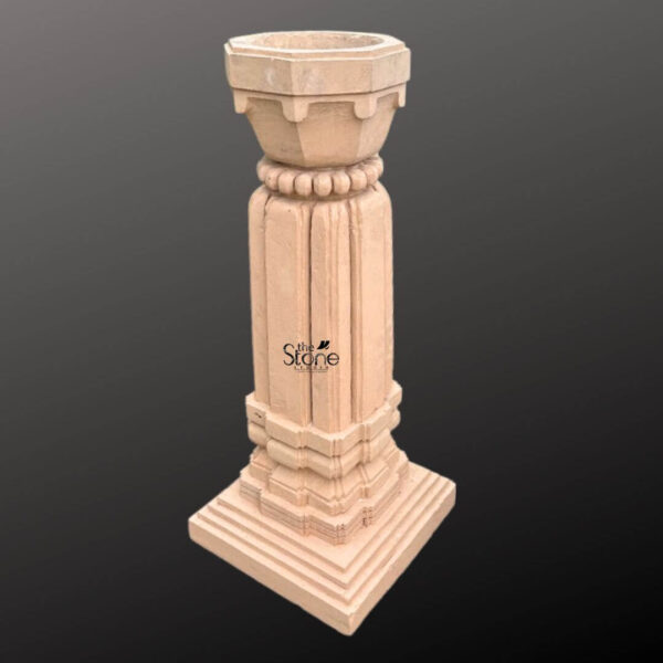 A three-quarter view photograph of a magnificent Outdoor Stone Tulsi Planter hand-carved from a single block of brown sandstone. Standing approximately 3 feet tall this traditional Indian planter is designed specifically for housing the sacred Tulsi plant. The entire structure rests upon a substantial stepped square base providing stability and a sense of grounded grandeur suitable for a garden or courtyard setting. The main body of the planter features a square column with a smooth hand-chiseled finish showcasing the natural texture and warm hues of the sandstone. The column is adorned with simple yet elegant vertical fluting creating clean lines that draw the eye upward. The four faces of the column exhibit a beautiful consistency in craftsmanship reflecting the skill and artistry of traditional stone carving techniques. Crowning the column is a beautifully shaped flared planter bowl designed to hold the sacred soil and plant. This upper section transitions from the square column to a circular form accented by a decorative band of carved bead-and-reel molding. The very top rim of the planter is finished with a simple clean edge. The overall form is a blend of architectural strength and delicate artistry making this sandstone planter a fitting and revered addition to any article discussing the significance and placement of a Tulsi plant in an outdoor space.