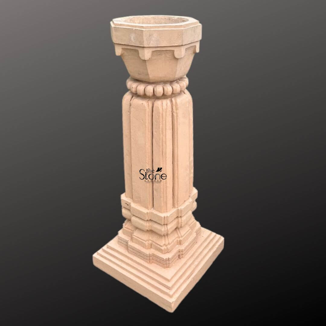 A three-quarter view photograph of a magnificent Outdoor Stone Tulsi Planter hand-carved from a single block of brown sandstone. Standing approximately 3 feet tall this traditional Indian planter is designed specifically for housing the sacred Tulsi plant. The entire structure rests upon a substantial stepped square base providing stability and a sense of grounded grandeur suitable for a garden or courtyard setting. The main body of the planter features a square column with a smooth hand-chiseled finish showcasing the natural texture and warm hues of the sandstone. The column is adorned with simple yet elegant vertical fluting creating clean lines that draw the eye upward. The four faces of the column exhibit a beautiful consistency in craftsmanship reflecting the skill and artistry of traditional stone carving techniques. Crowning the column is a beautifully shaped flared planter bowl designed to hold the sacred soil and plant. This upper section transitions from the square column to a circular form accented by a decorative band of carved bead-and-reel molding. The very top rim of the planter is finished with a simple clean edge. The overall form is a blend of architectural strength and delicate artistry making this sandstone planter a fitting and revered addition to any article discussing the significance and placement of a Tulsi plant in an outdoor space.