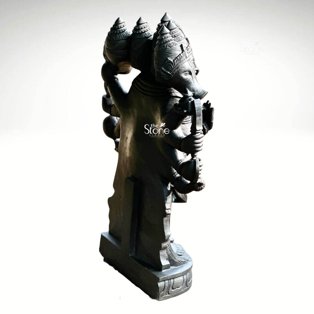 A three-quarter rear view studio photograph set against a plain light grey background displays a magnificent 3.5-foot Panchamukha Anjaneya Stone Vigraham. This intricately hand-carved statue is made from a single block of deep dark blackstone renowned for its durability and fine grain. The statue depicts the five-faced form of Lord Hanuman known as Panchamukhi Hanuman standing firmly on a multi-tiered, ornate circular base. From this rear-three-quarter angle the complexity of the carving is evident with the multiple heads arranged in a tiered fashion rising above the broad shoulders. The central head is the most prominent adorned with an elaborate multi-tiered kiritamukuta that is deeply carved with intricate patterns and a central motif. The view highlights the powerful, broad back and shoulders of the deity covered with traditional South Indian carvings of necklaces armlets wristlets a sacred thread and a detailed dhoti belt all chiseled with precision into the dark stone. The multiple arms, totaling ten, are arranged symmetrically, holding various divine attributes. From this perspective the left-side arms are clearly visible each holding an object such as a conch discus sword shield noose and hook rendered with exquisite detail. The right-side arms are seen in profile mirroring the arrangement with objects like a mace mountain and vessel. The fine texture of the blackstone enhances the depth and shadow of the carvings highlighting the complex iconography of this formidable yet benevolent form of Hanuman. This Panchamukha Anjaneya Stone Vigraham is an exceptional example of traditional Indian temple sculpture suitable for sanctum placement or as a grand focal point in a home shrine or spiritual space. The 3.5-foot height makes it a commanding presence embodying the combined power of the five deities represented by the faces. The blackstone material gives the statue a timeless, ancient feel ensuring it will be a cherished piece for generations. The statue's detailed iconography from the expressions on the five faces to the specific items held in the ten hands makes it a significant subject for study and devotion. The expert craftsmanship transforms the raw stone into a vibrant sacred form.