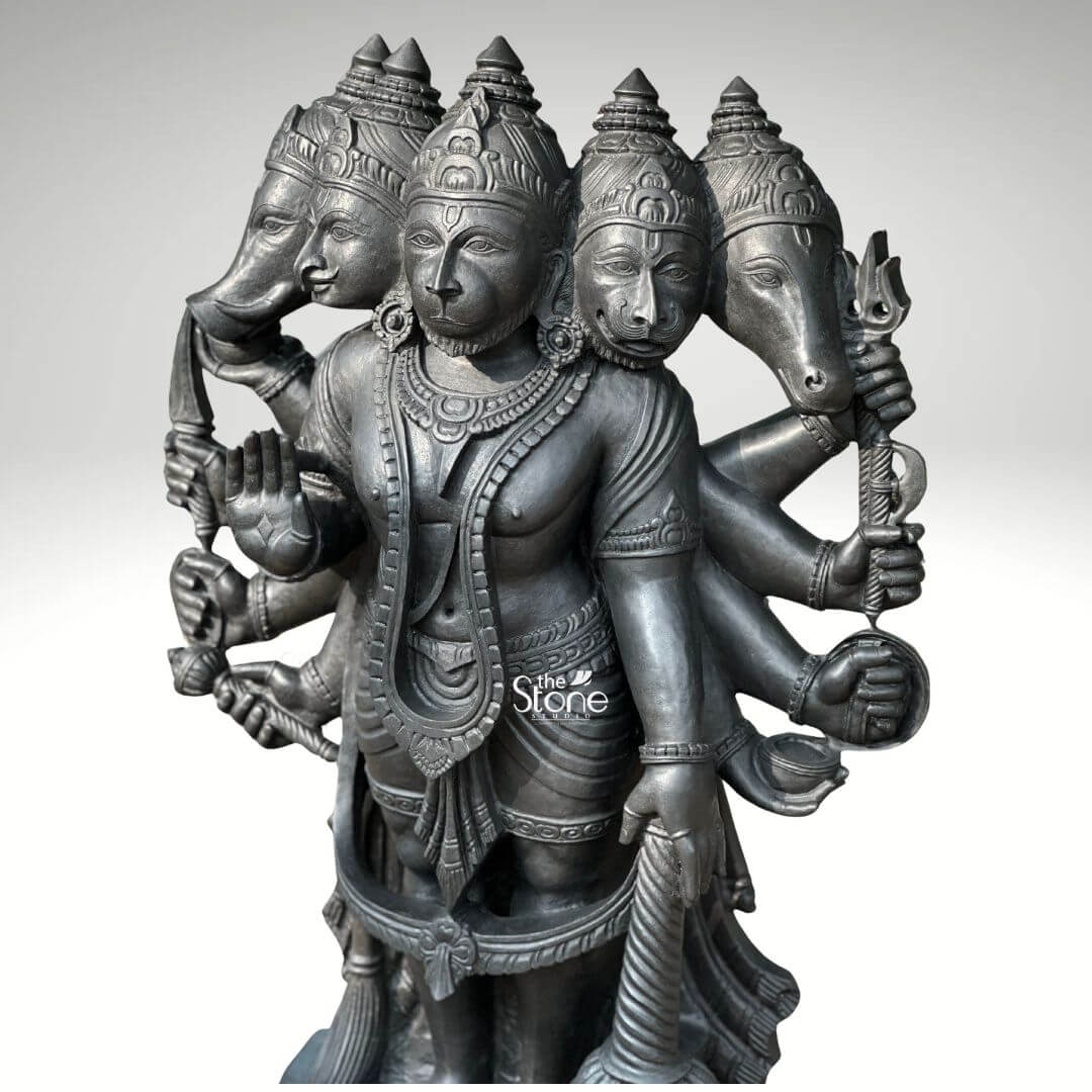 A full-view studio photograph set against a plain light grey background displays a magnificent 3.5-foot Panchamukha Anjaneya Swamy Stone Statue. This intricately hand-carved Vigraham is made from a single block of deep dark blackstone renowned for its durability and fine grain. The statue depicts the five-faced form of Lord Hanuman known as Panchamukhi Hanuman standing firmly on a multi-tiered, ornate circular base. The central face is that of Hanuman himself looking forward with a serene protective expression adorned with traditional jewelry and a crown. Radiating outwards from the central head are four other divine faces: to the east the boar face to the south the lion face to the west the eagle face and to the north the horse face. The statue is multi-arme totaling ten arms that hold various divine attributes each rendered with exquisite detail. The main central right hand is raised in the Abhaya mudra while the main central left hand holds a mace that rests on the base. The other eight hands hold a conch discus sword shield noose hook a mountain and a vessel. The deity is adorned with elaborate traditional South Indian carvings of necklaces armlets wristlets a sacred thread and a detailed dhoti all chiseled with precision into the dark stone. The fine texture of the blackstone enhances the depth and shadow of the carvings highlighting the complex iconography of this formidable yet benevolent form of Hanuman. This Panchamukha Anjaneya Swamy Stone Statue is an exceptional example of traditional Indian temple sculpture suitable for sanctum placement or as a grand focal point in a home shrine or spiritual space. The 3.5-foot height makes it a commanding presence embodying the combined power of the five deities represented by the faces. The blackstone material gives the statue a timeless ancient feel ensuring it will be a cherished piece for generations. The statue's detailed iconography from the expressions on the five faces to the specific items held in the ten hands makes it a significant subject for study and devotion. The expert craftsmanship transforms the raw stone into a vibrant sacred form.