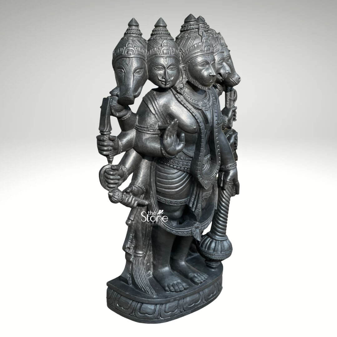A full-view studio photograph set against a plain light grey background displays a magnificent 3.5-foot Panchamukha Anjaneya Swamy Vigraham for Temple. This intricately hand-carved statue is made from a single block of deep dark blackstone renowned for its durability and fine grain. The statue depicts the five-faced form of Lord Hanuman known as Panchamukhi Hanuman standing firmly on a multi-tiered ornate circular base that is ready for temple installation. The central face is that of Hanuman himself looking forward with a serene protective expression adorned with traditional jewelry and a crown. Radiating outwards from the central head are four other divine faces: to the east the boar face to the south the lion face to the west the eagle face and to the north the horse face. The statue is multi-armed totaling ten arms that hold various divine attributes each rendered with exquisite detail. The main central right hand is raised in the Abhaya mudra while the main central left hand holds a mace that rests on the base. The other eight hands hold a conch discus sword shield noose hook a mountain and a vessel. The deity is adorned with elaborate, traditional South Indian carvings of necklaces armlets wristlets a sacred thread and a detailed dhoti all chiseled with precision into the dark stone. The fine texture of the blackstone enhances the depth and shadow of the carvings highlighting the complex iconography of this formidable yet benevolent form of Hanuman. This Panchamukha Anjaneya Swamy Vigraham is specifically designed for temple sanctum placement, embodying the combined power of the five deities represented by the faces. The 3.5-foot height makes it a commanding presence, suitable for daily puja and darshan. The blackstone material gives the statue a timeless, ancient feel, ensuring it will be a revered centerpiece for generations. The statue's detailed iconography, from the expressions on the five faces to the specific items held in the ten hands, makes it a significant subject for study and devotion. The expert craftsmanship transforms the raw stone into a vibrant, sacred form, fulfilling the requirements for a traditional temple vigraham.