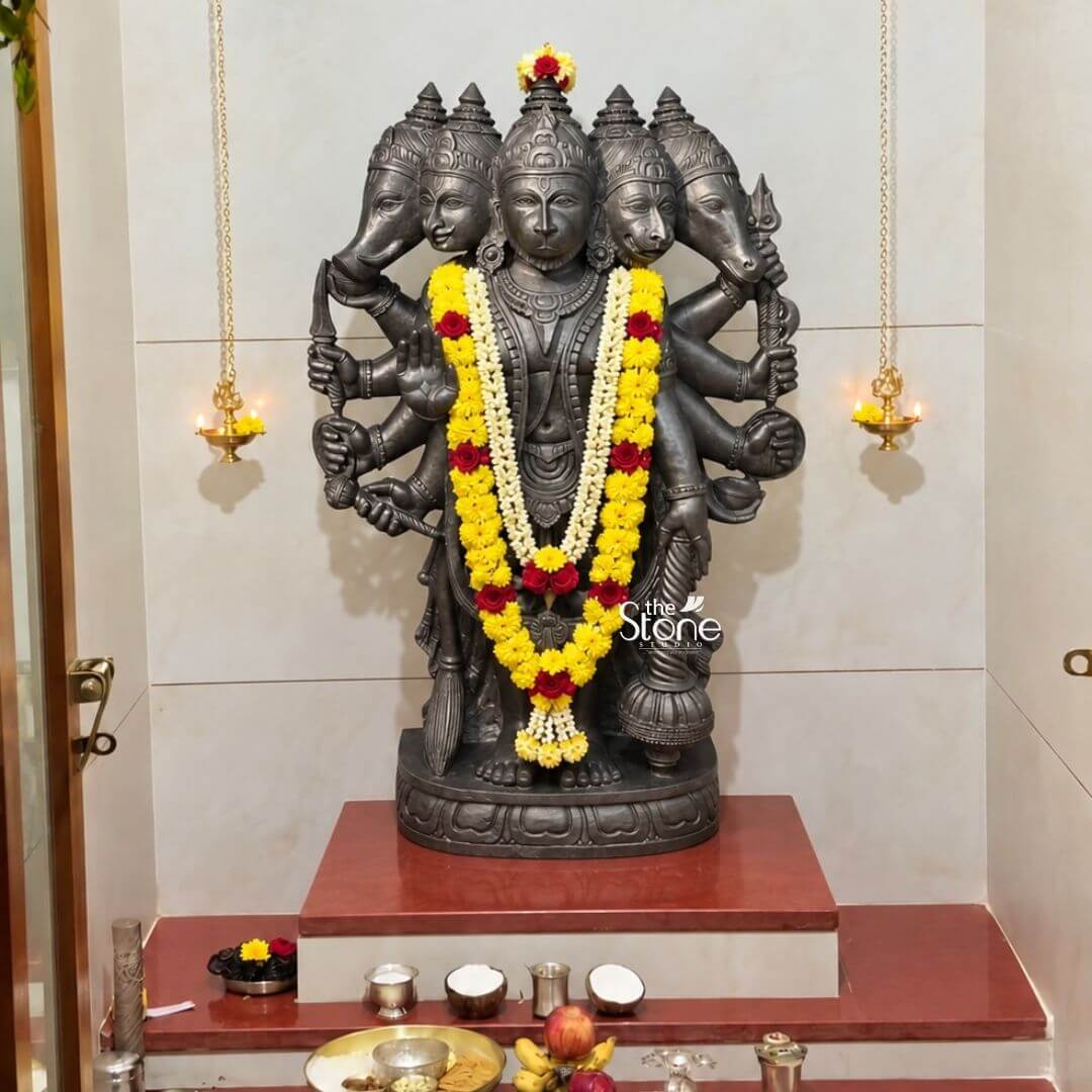 A centered medium shot captures a beautifully consecrated home shrine centered on a magnificent 3.5-foot Panchamukha Hanuman Black Stone Vigraham. The statue hand-carved from a single block of deep dark blackstone depicts the five-faced form of Lord Hanuman known as Panchamukhi Hanuman standing firmly on a multi-tiered ornate circular base. The central face is that of Hanuman himself looking forward with a serene protective expression. Radiating outwards from the central head are four other divine faces to the east the boar face to the south the lion face to the west the eagle face and to the north the horse face. The detailed iconography chiseled with precision into the dark stone includes ten arms holding various attributes such as a conc discus sword shield noose hook mountain and vessel with the main right hand raised in blessing. The blackstone Vigraham rests upon a polished reddish-brown marble pedestal, elevated on a white platform within a recessed wall niche. The shrine is richly decorated for worship. The statue is adorned with a prominent thick double-strand garland of fresh yellow marigolds and red roses along with a smaller white flower garland draped across the chest all accented with small pink rosebuds. Two traditional brass oil lamps with burning wicks are suspended by chains from the ceiling of the alcove casting a warm flickering golden glow upon the deity. The pedestal is adorned with puja items small polished silver bowls containing kumkum and haldi a cluster of small coconuts, and a decorative brass plate in the foreground holding fruits like bananas and pomegranates as well as other ritual offerings. The overall atmosphere of the shrine is one of profound peace and spiritual devotion making it a fitting focal point within a traditional Indian home. The dark stone of the Hanuman Vigraham provides a powerful contrast against the light cream-colored tiled wall while the fresh yellow red and white flowers add a vibrant touch of color and life. The arrangement demonstrates a classic home puja setup showcasing the 3.5-foot blackstone statue as the central object of worship. The soft warm lighting from the hanging brass lamps emphasizes the smooth contours and detailed carvings of the deity creating a sacred space for daily prayer and reflection. The image conveys the beauty and sanctity of traditional Indian stone artistry in a grand scale.